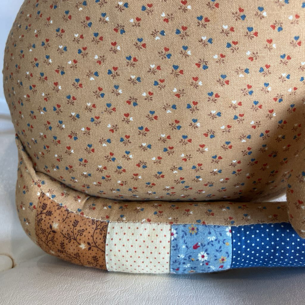 A close-up of a handmade fabric cushion or chair backrest featuring a beige linen-like base printed with a small red, blue, and white floral and heart pattern, with a patchwork trim of brown, cream, light blue, and dark blue fabrics.