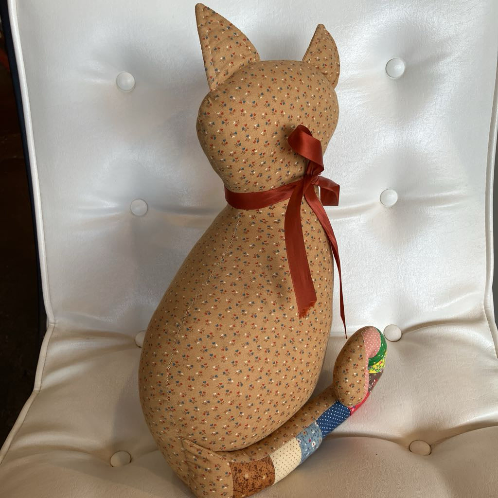 A handmade fabric cat-shaped cushion or decorative figure sits on a white tufted chair. The figure is made from tan fabric with a small blue and orange floral pattern, has a red satin ribbon tied around its neck, and a multicolored patchwork tail.
