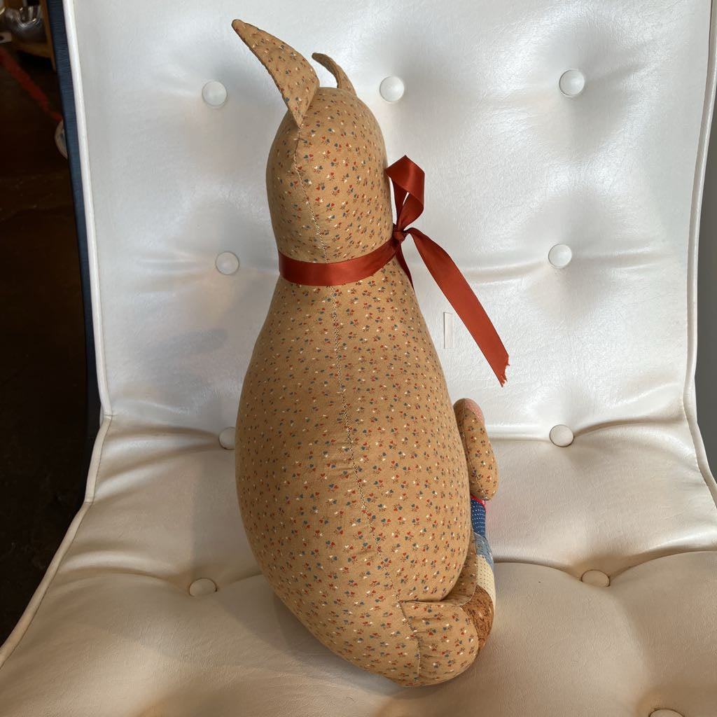 A handmade fabric dog doll with a tan base, tiny multicolor floral pattern, and a red ribbon bow, sitting on a white tufted chair.