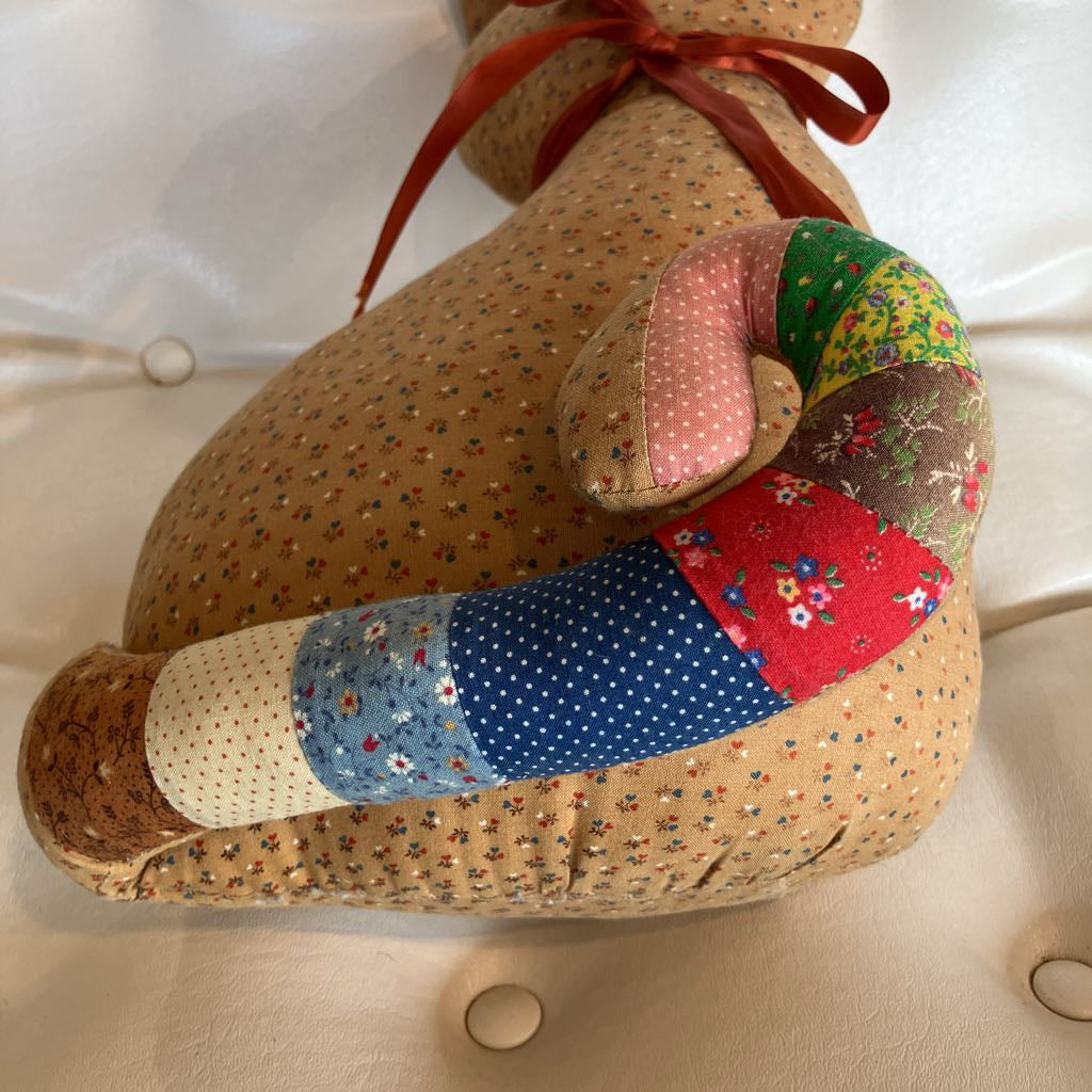 A handmade stuffed animal or cushion-like plush toy with a tan floral fabric body, a curved patchwork tail made of pink, green, brown, blue, red, and cream fabrics, and a red ribbon tied at the neck, photographed on a white tufted surface.