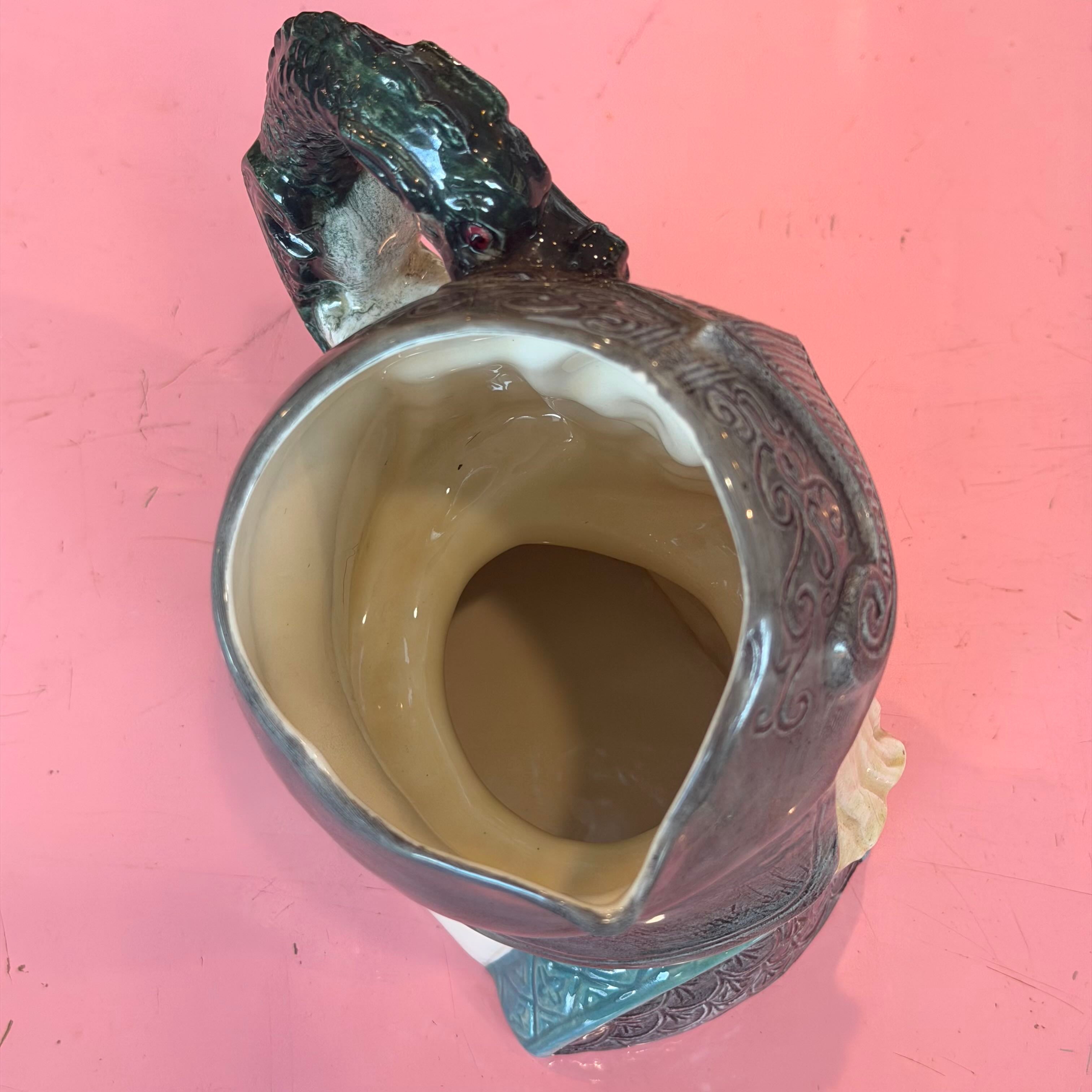 A ceramic pitcher shaped like a frog, with a glossy dark green head and a speckled gray-green body, photographed from above on a pink background.