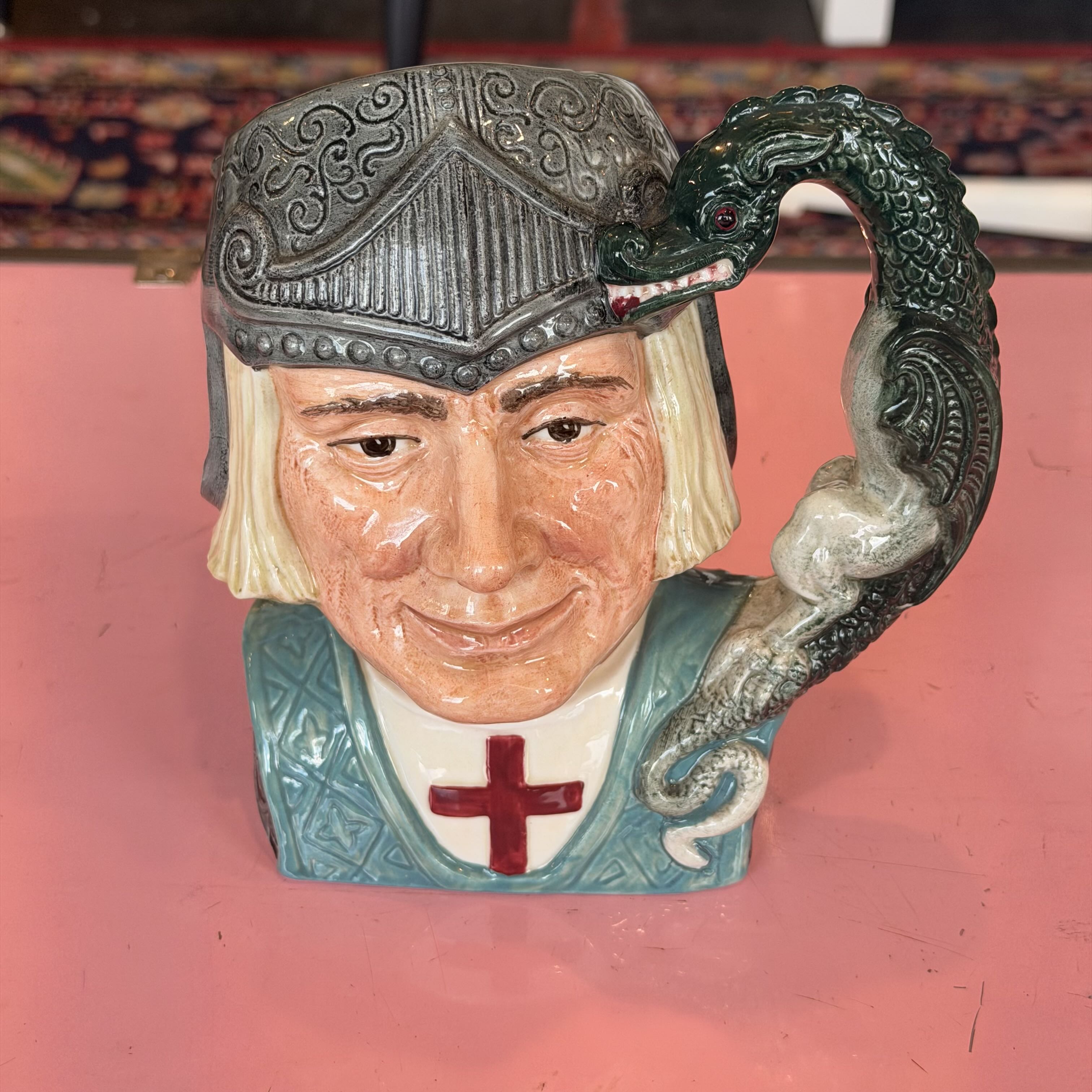 A ceramic Toby jug shaped like a knight's bust with a dark green dragon handle wrapping around the side, set against a pink background.
