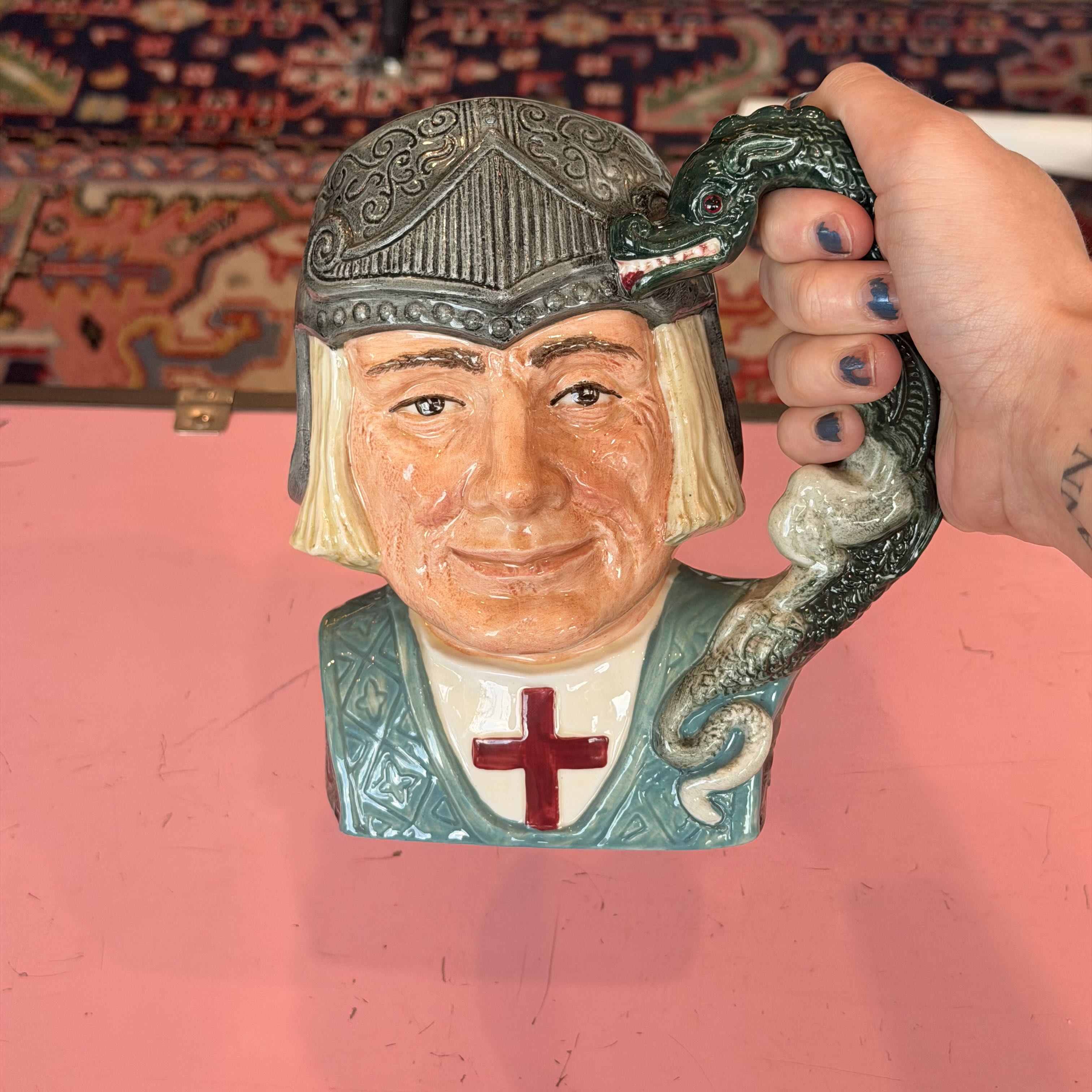 A ceramic Toby-style mug shaped like a medieval knight's bust with a green dragon handle, held by a hand against a pink tabletop and patterned rug background.