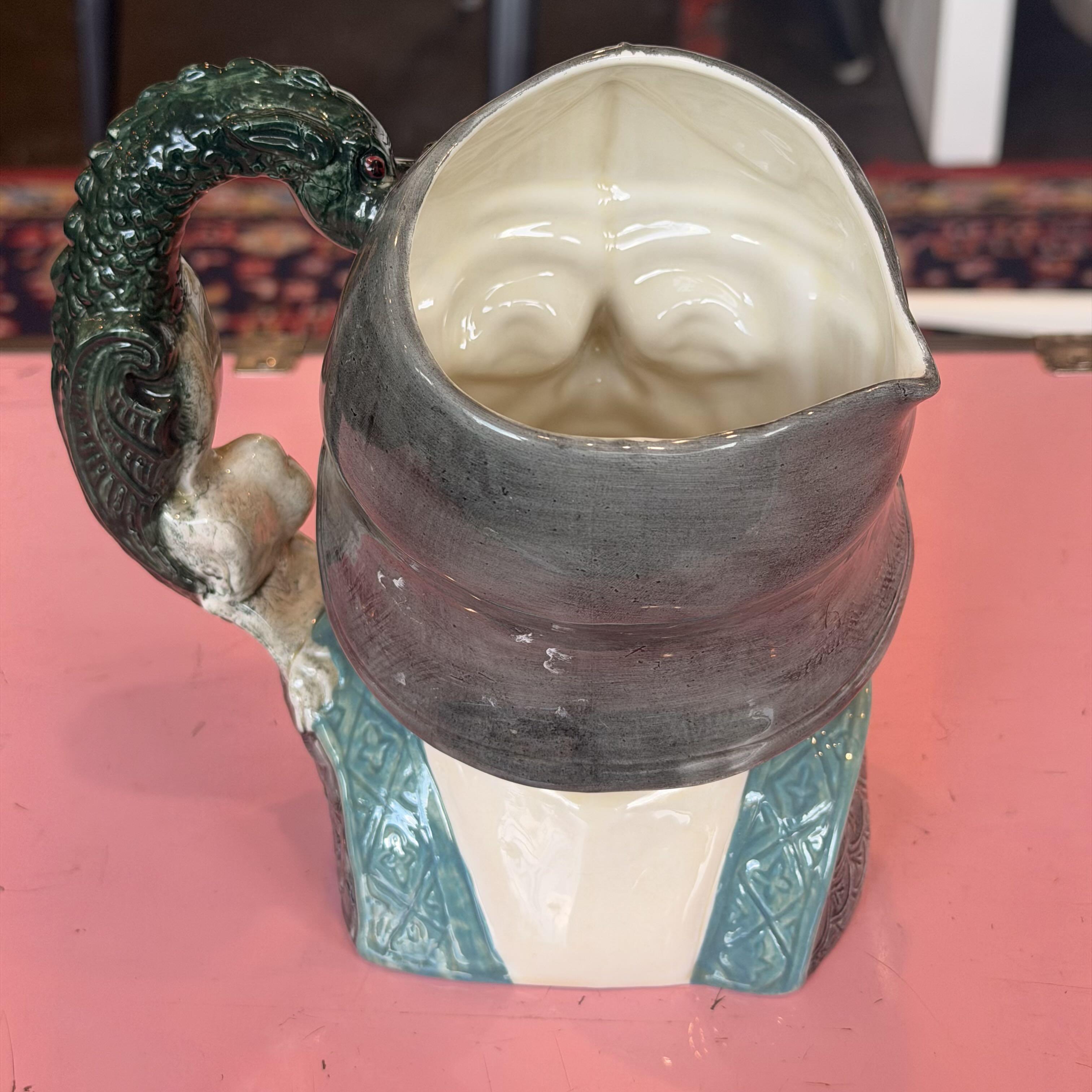 A ceramic pitcher shaped like a stylized face wearing a dark gray hat, with a green dragon-like handle and turquoise patterned base, set against a pink background.