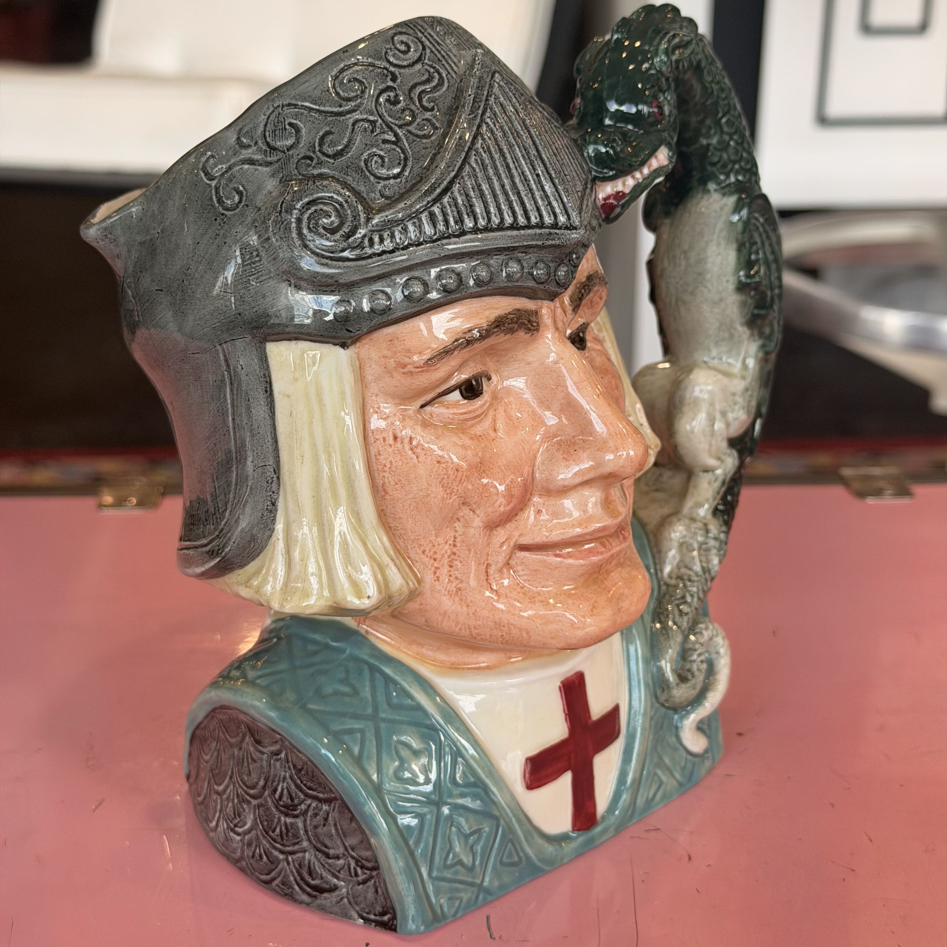 A ceramic novelty jug shaped like a medieval knight's bust holding a dragon on its shoulder, displayed on a pink surface.