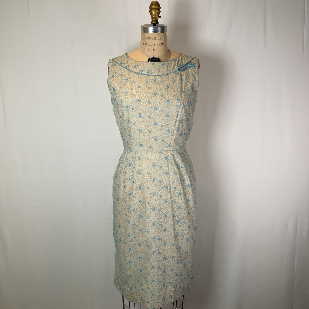 A sleeveless knee-length dress with a gathered waist, featuring a light beige and white floral pattern with blue accents, displayed on a vintage dress form mannequin against a white fabric backdrop.