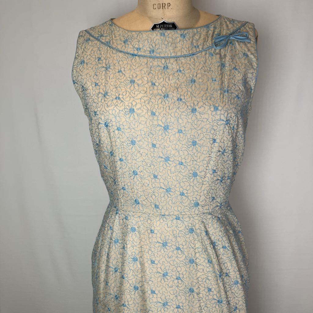 A vintage sleeveless dress displayed on a mannequin torso against a plain light gray background. The dress has a light beige or cream base with a blue floral lace-like pattern and a matching blue bow detail at the neckline.