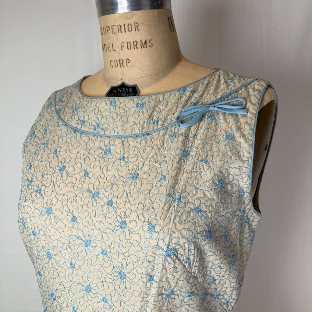 A sleeveless cream-colored dress form bust with a light blue floral pattern and a matching blue bow detail on the neckline, displayed on a vintage wooden dress form.