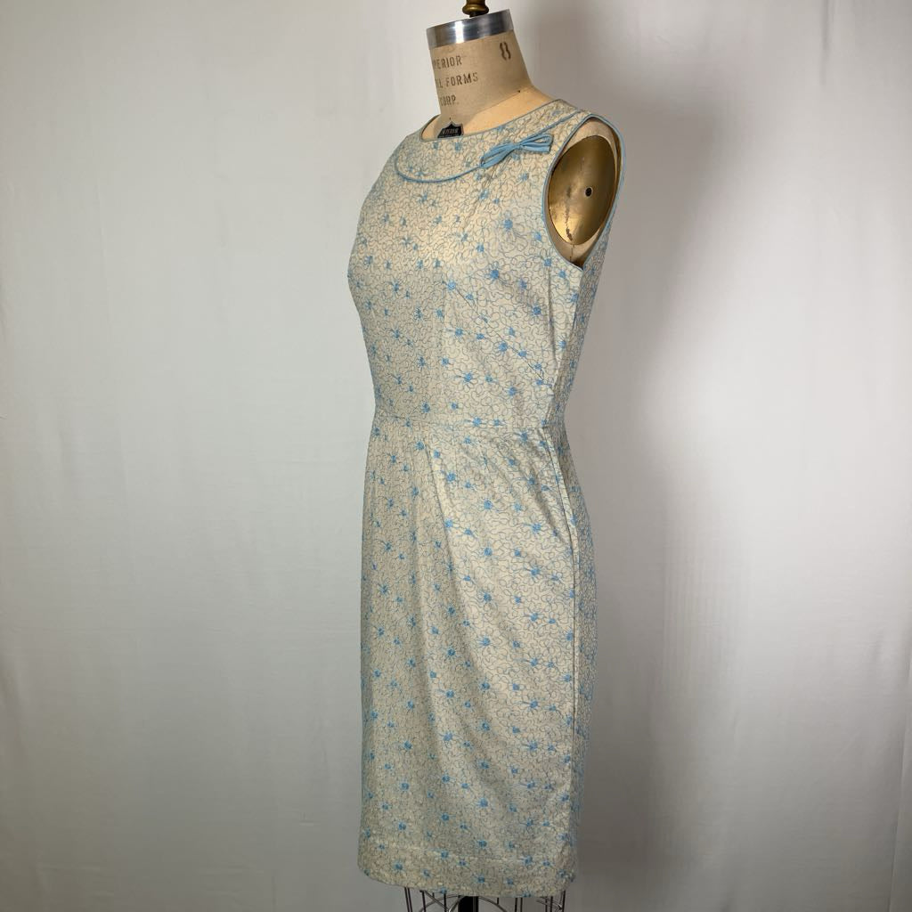 A sleeveless knee-length dress with a small blue floral print on a light beige or off-white base, displayed on a vintage dress form mannequin against a white fabric backdrop.