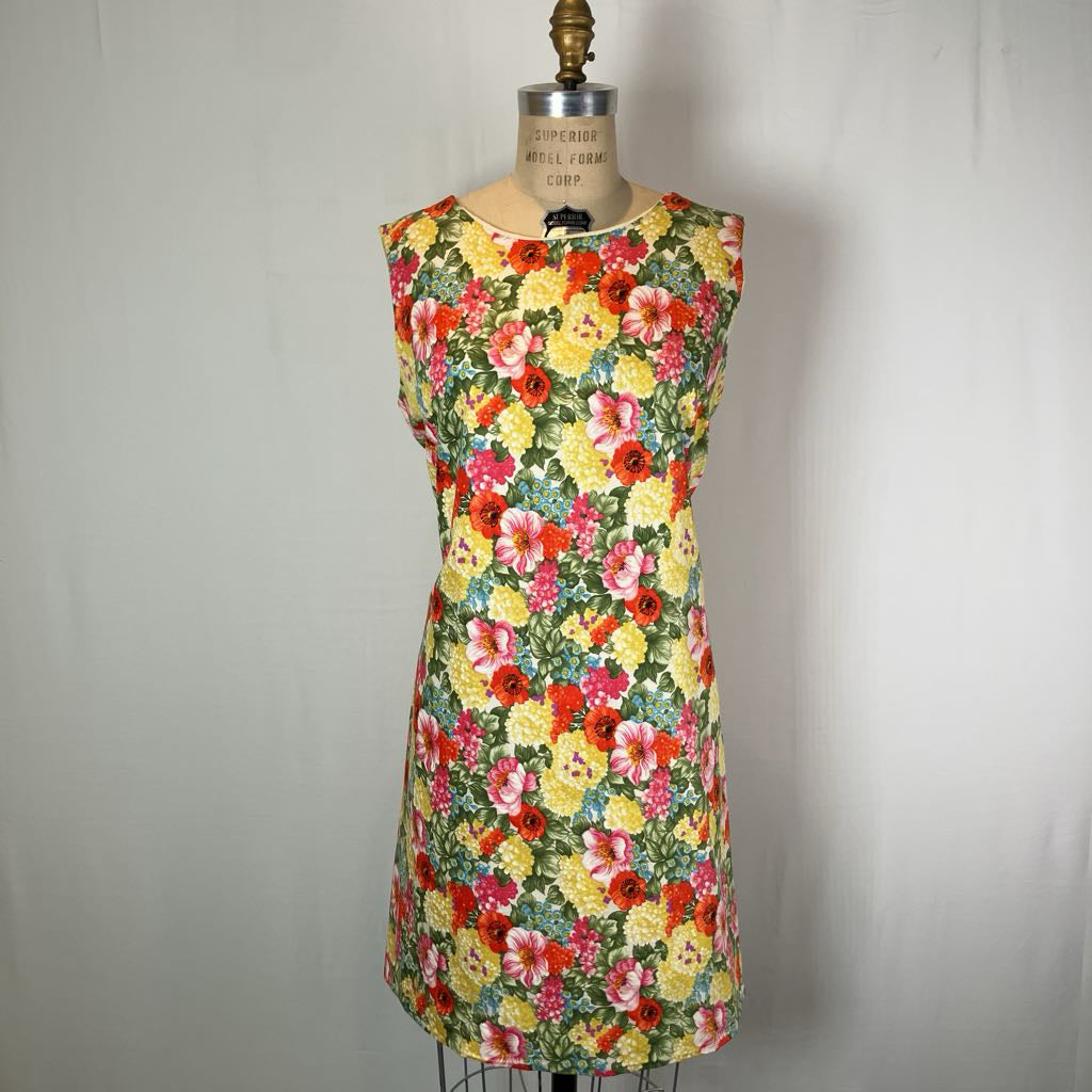 A sleeveless A-line dress with a vibrant multicolor floral print, displayed on a vintage dress form mannequin against a neutral off-white fabric backdrop.