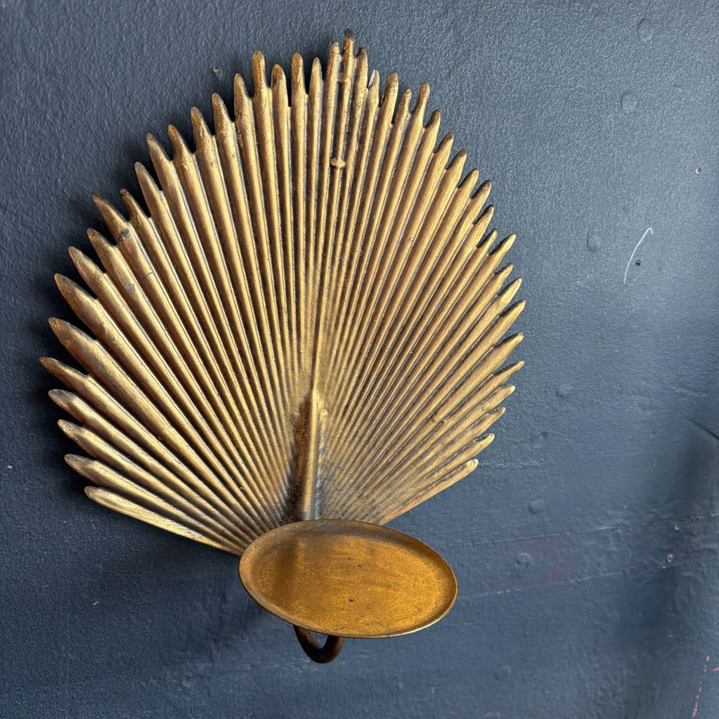 A vintage brass wall sconce shaped like a fan or palm leaf, mounted on a dark textured wall.