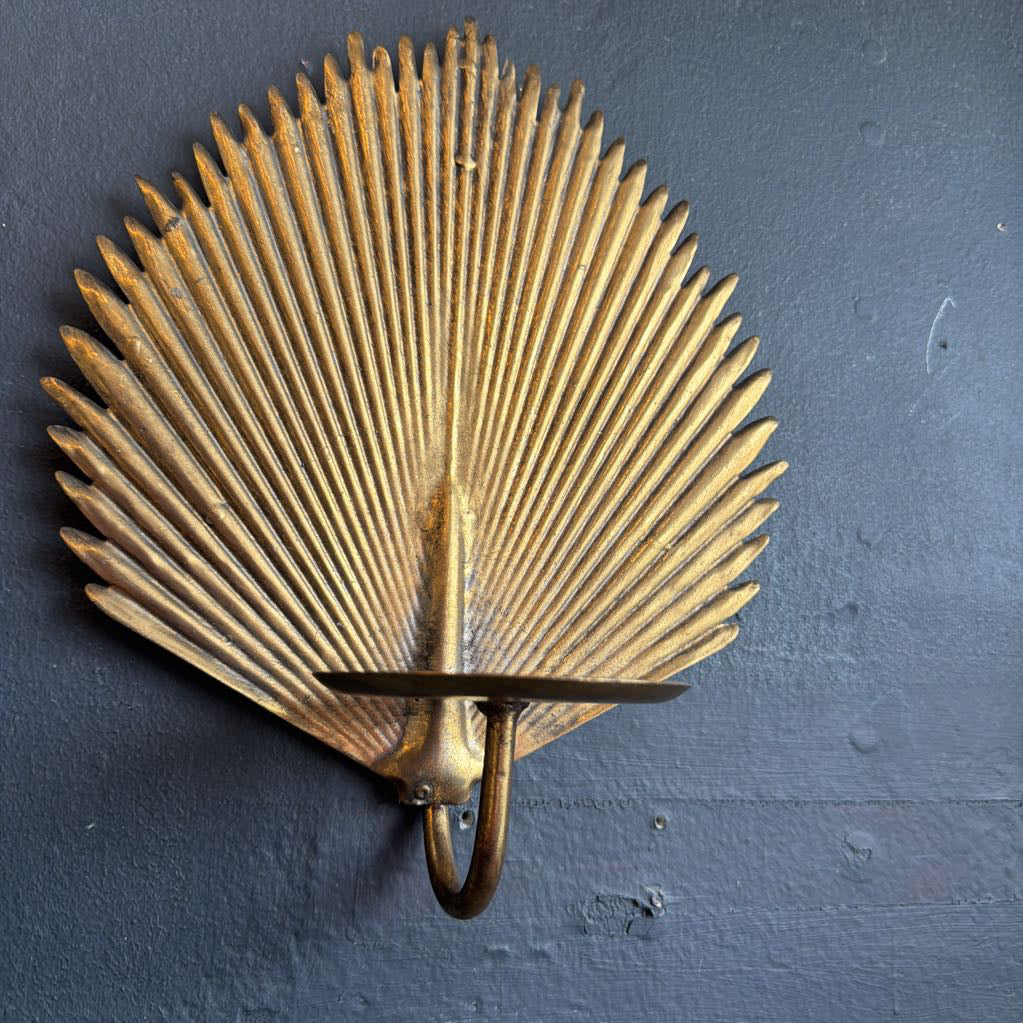 A vintage-style metal wall sconce shaped like a fan or scallop, with a warm gold and bronze finish, mounted against a dark textured wall.