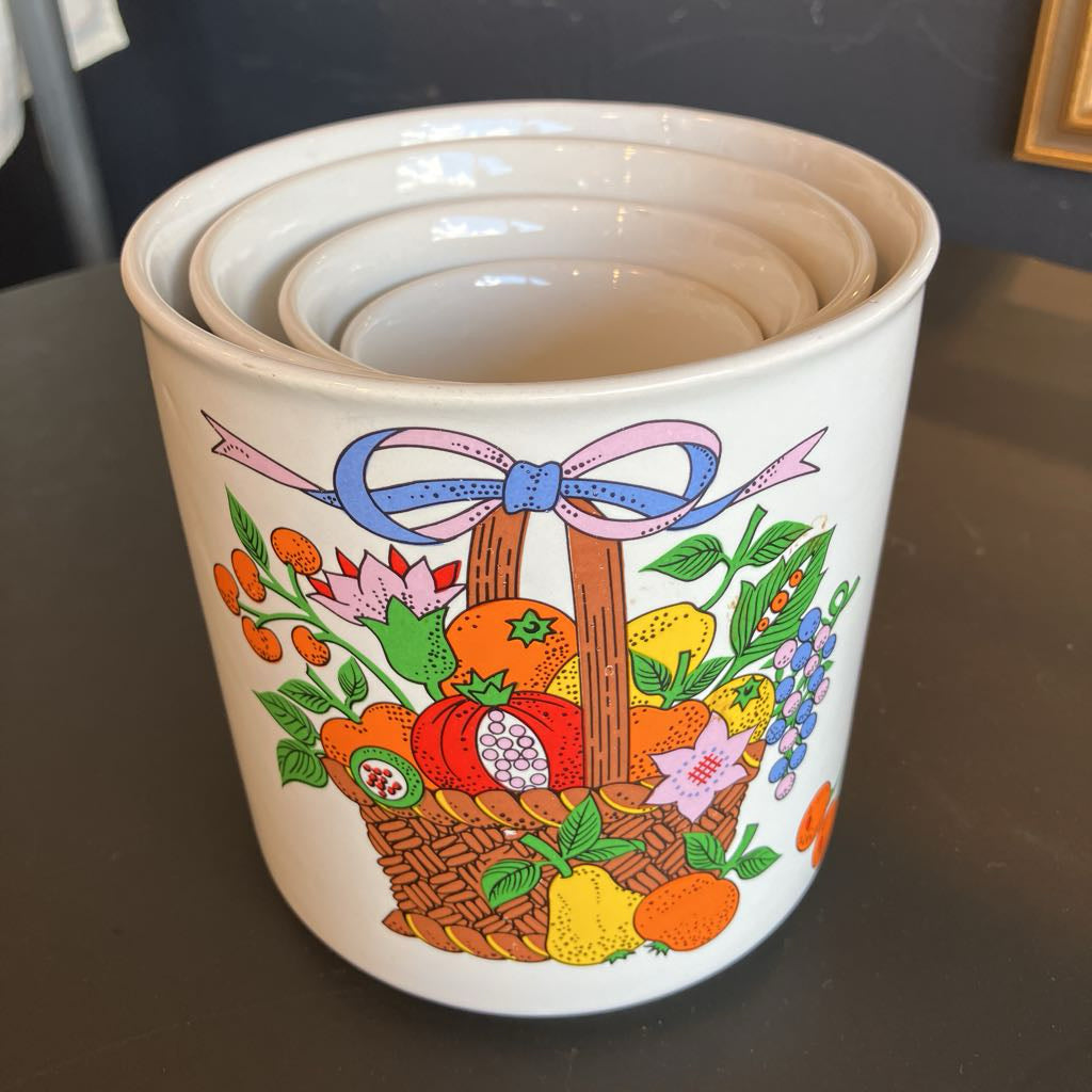 A white ceramic nesting canister with a colorful fruit and flower basket illustration, photographed on a dark tabletop.