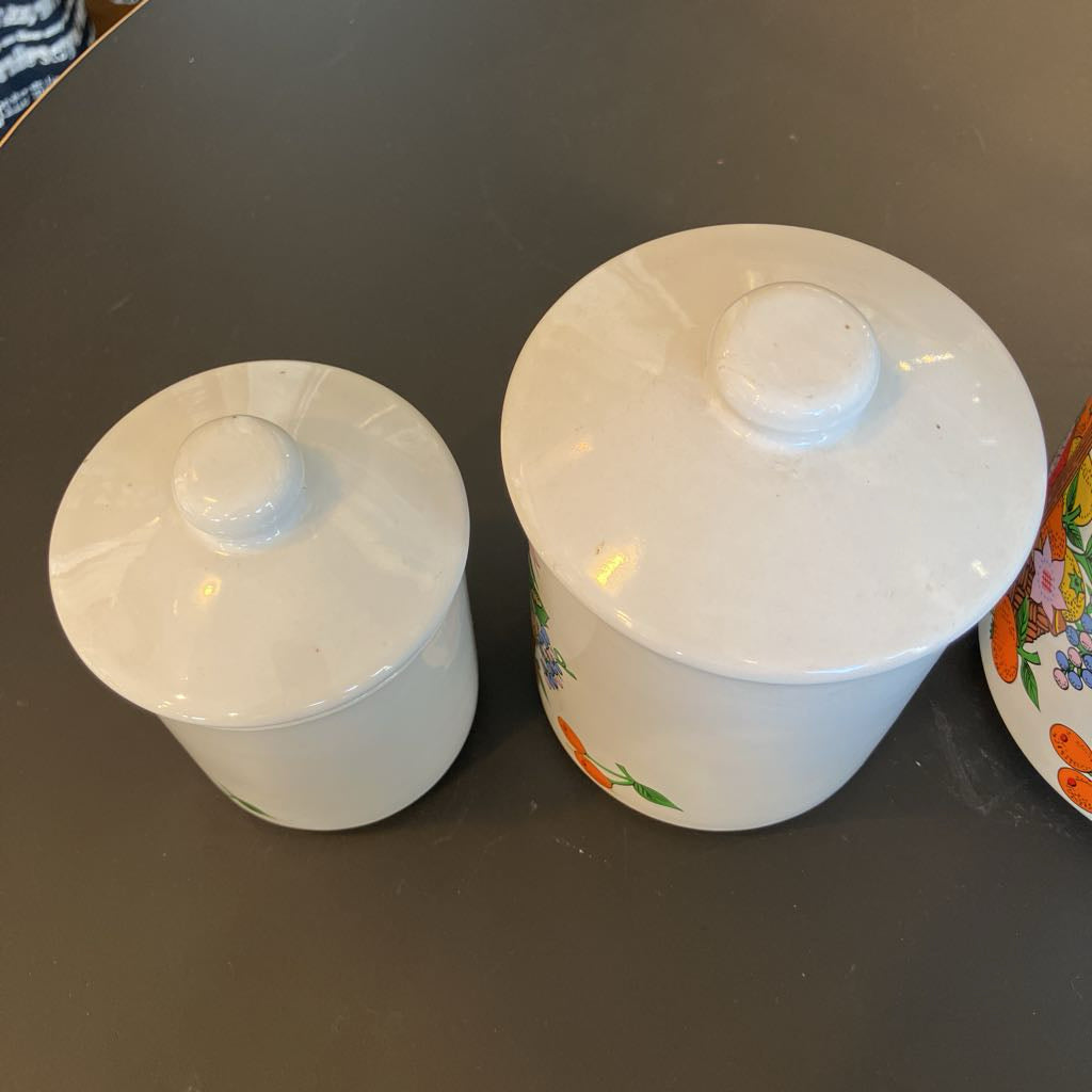 Two white ceramic lidded containers with colorful floral and fruit illustrations, shown on a dark tabletop.