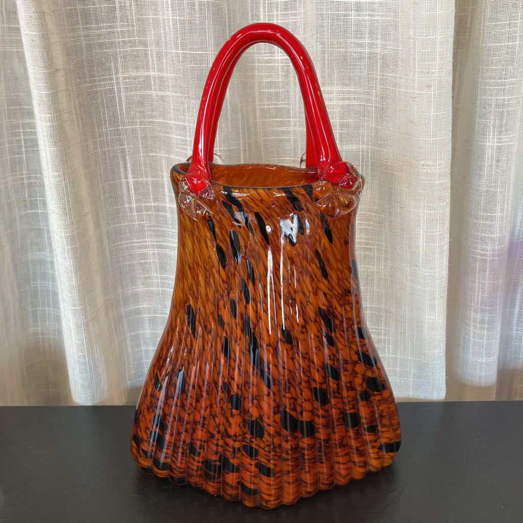 A vintage-style glass handbag with a pleated body in orange-brown and black swirl patterns, featuring a glossy red top handle, set against a neutral curtain background.