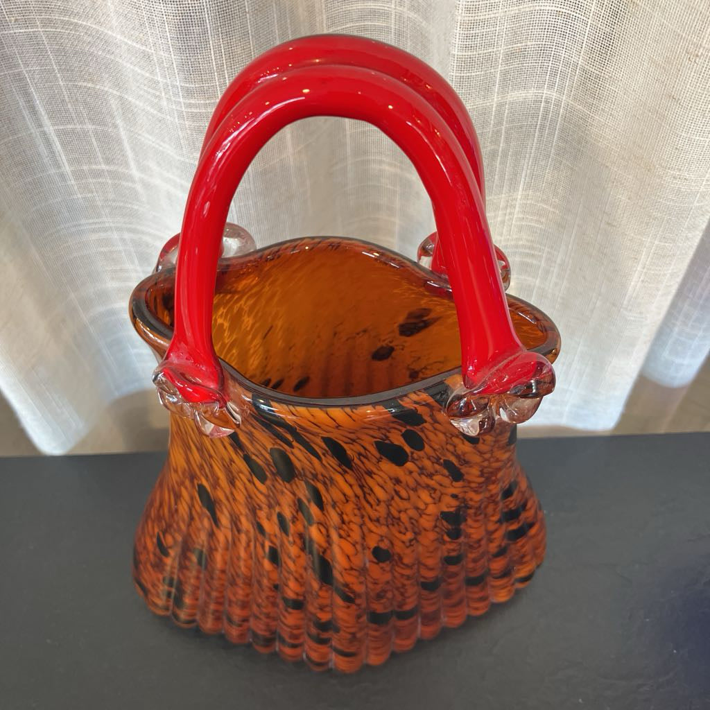An amber and orange glass basket with a red handle, featuring a scalloped lower edge and black speckled swirls, set against a white curtain background.