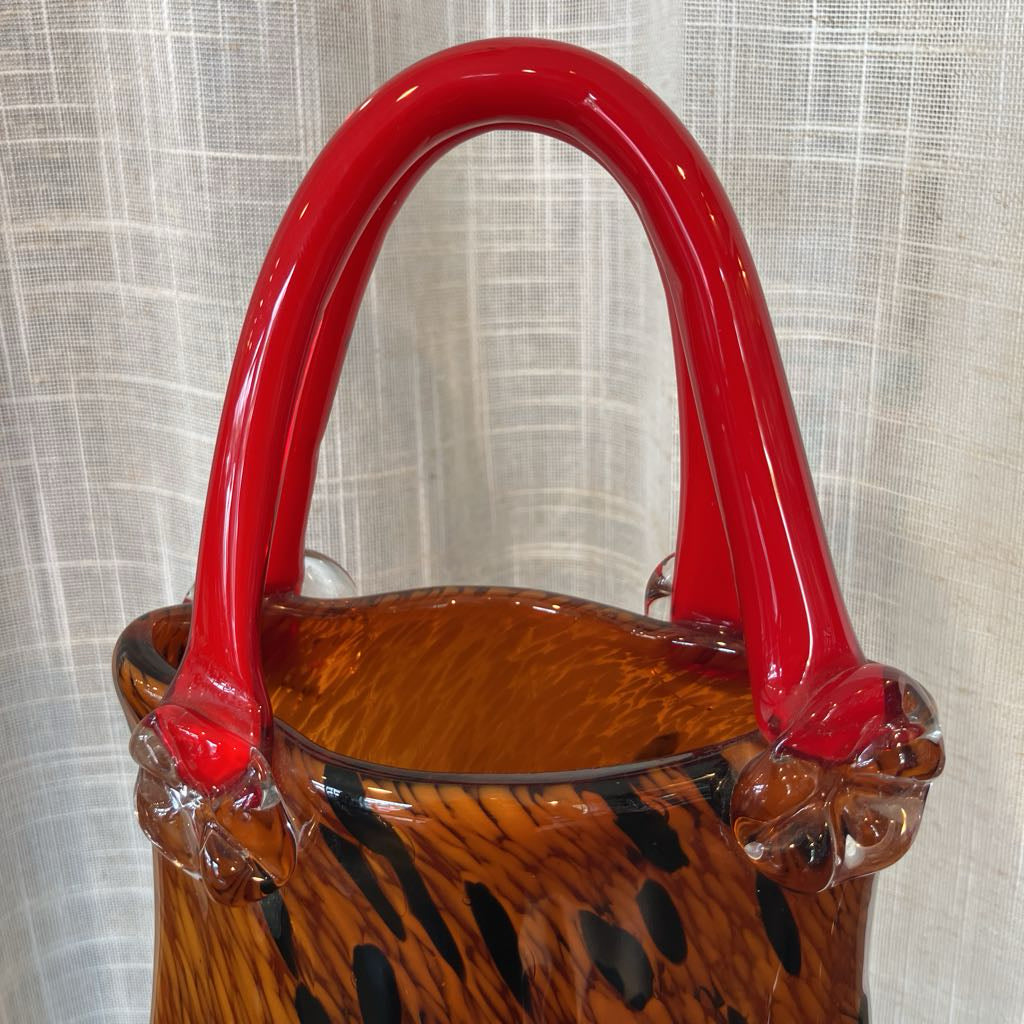 A close-up of a hand-blown glass vase or decorative container with a glossy red handle and a brown amber body featuring black leopard-like spots, set against a light textured curtain background.