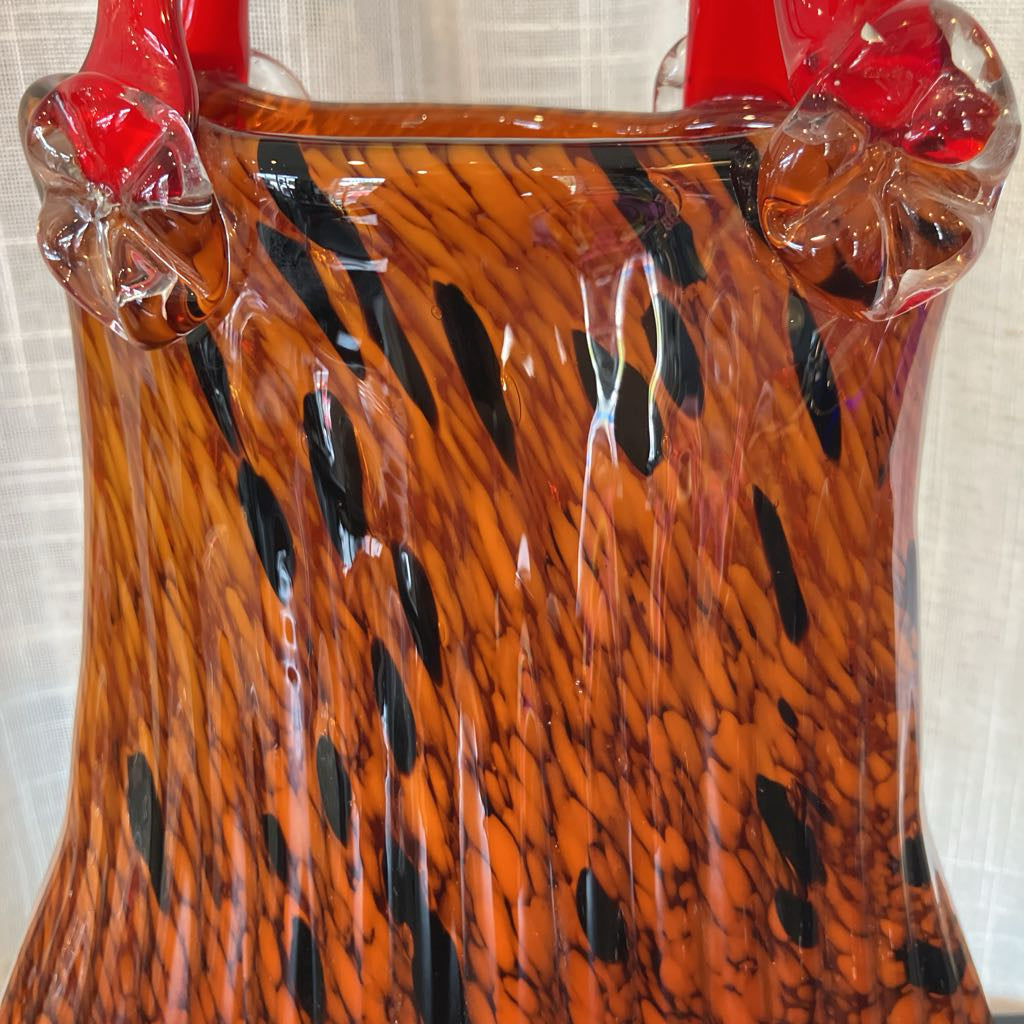 A close-up of a hand-blown glass vase featuring a vibrant orange and black tiger-stripe pattern, with glossy red handles and clear textured glass accents at the top.