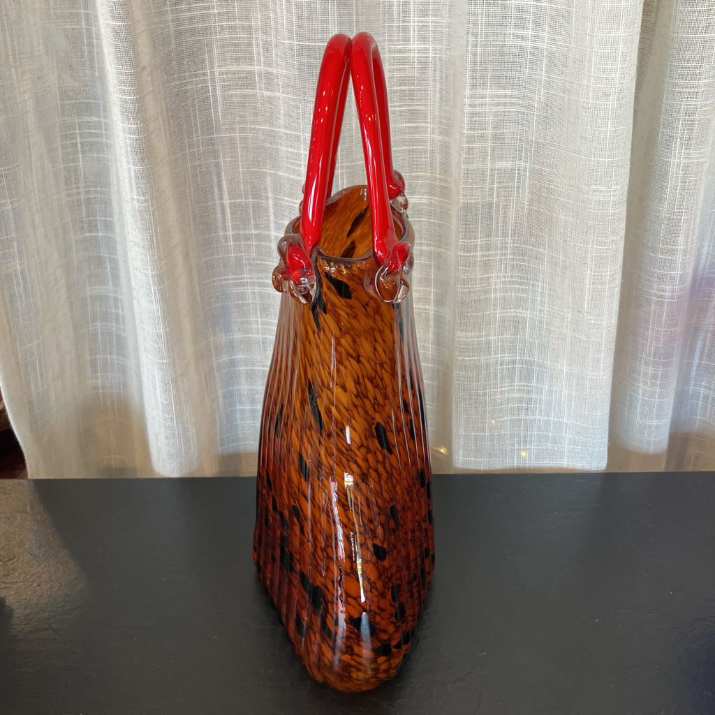 A vintage-style glass handbag with a glossy amber-brown body featuring black speckled patterns and vibrant red handles, set against a neutral curtain background.