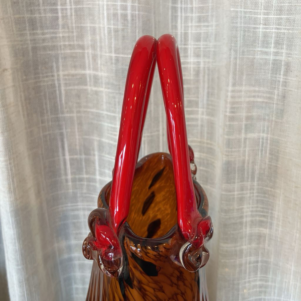 A close-up of a hand-blown glass vase with glossy red handles and a brown amber glass body, set against a white textured curtain background.