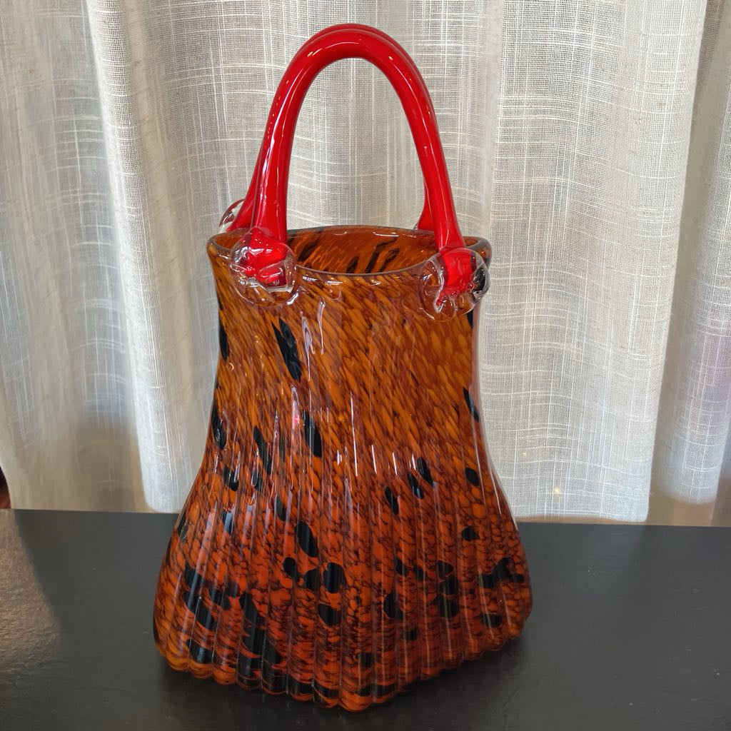 A vintage-style glass handbag with a textured orange and black patterned body and glossy red handles, shown on a dark tabletop in front of a light beige curtain.