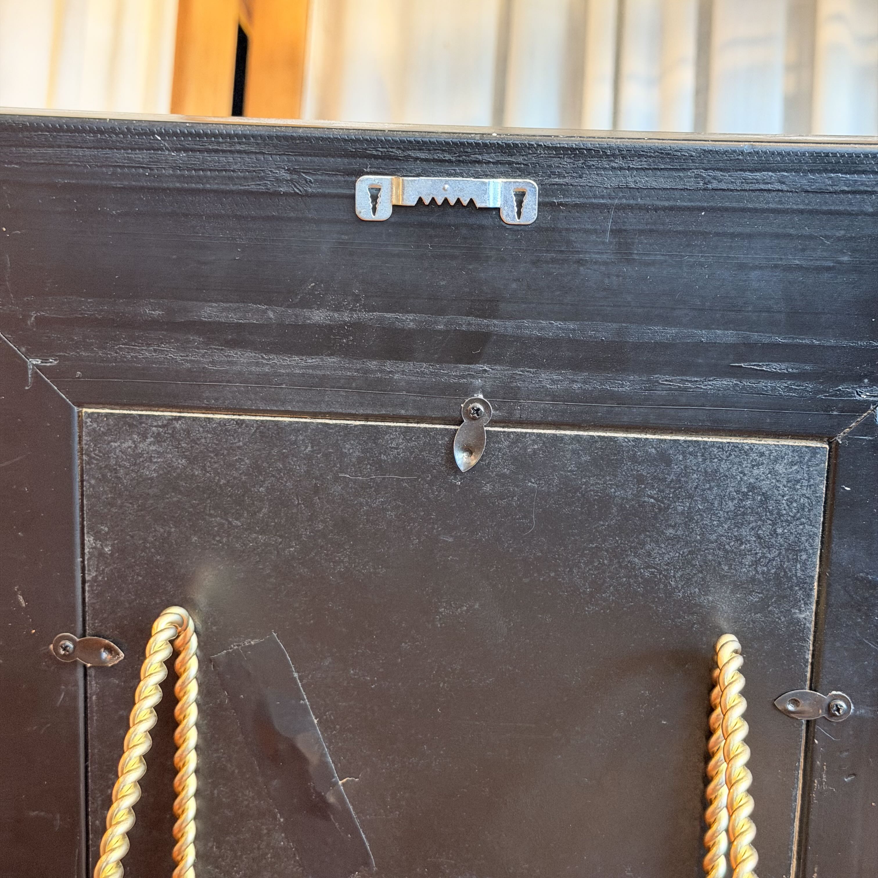 Close-up of the back of a black framed picture frame, showing the mounting hardware, a small metal slot near the top, and two gold-toned hanging ropes on the sides.