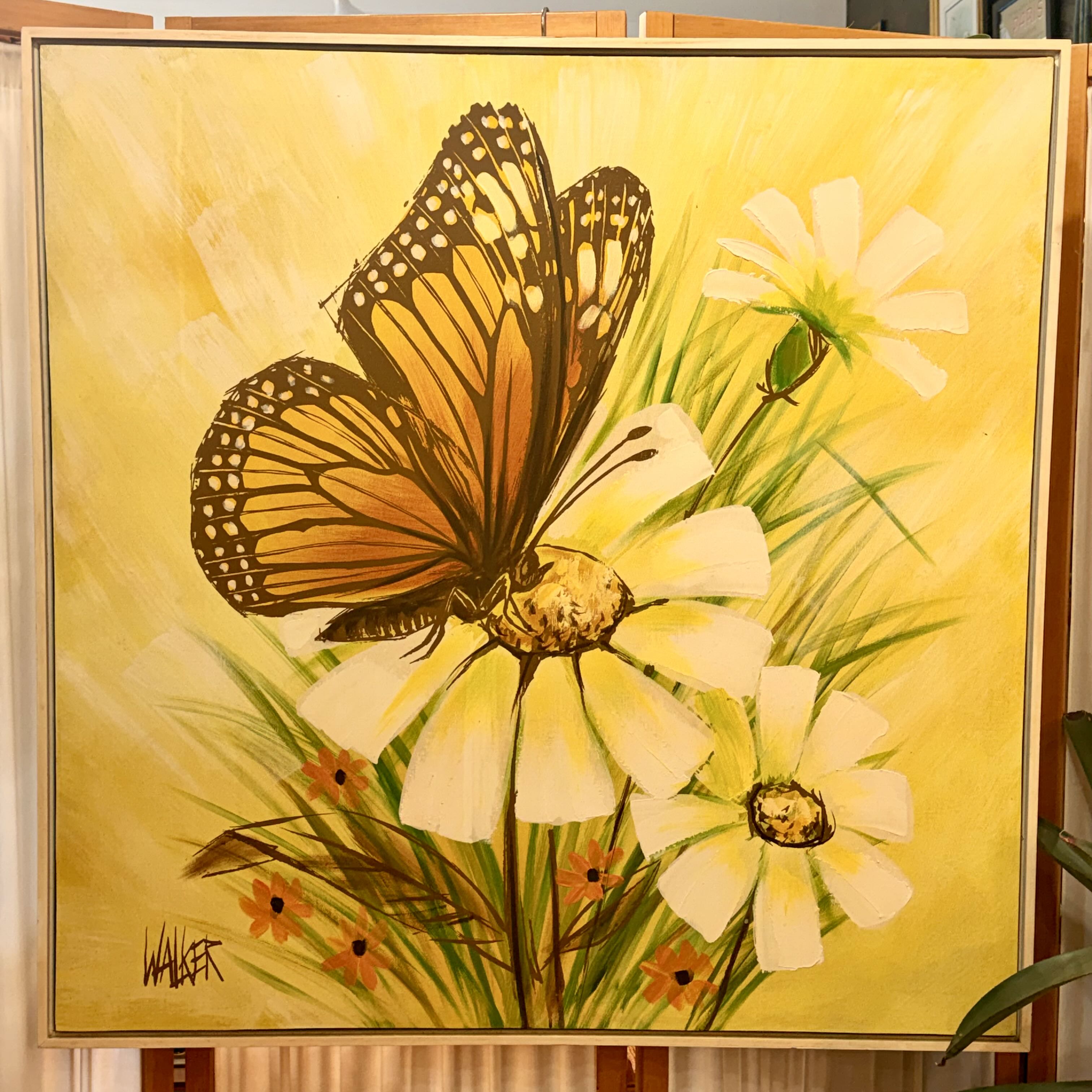 A framed painting on an easel depicting a large orange and black butterfly perched on white flowers with green stems against a bright yellow background. The artwork is signed by the artist in the lower left corner.