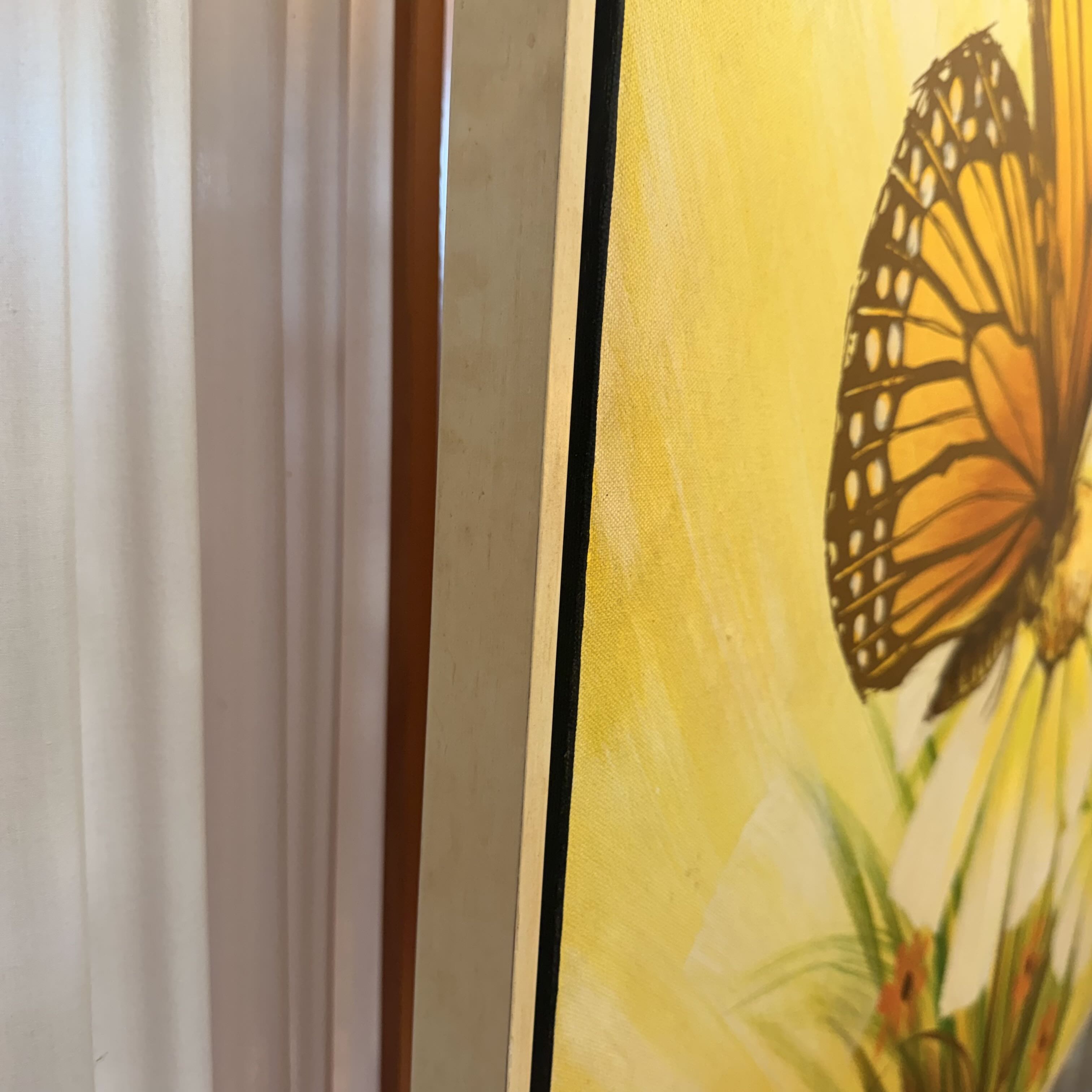 A close-up of a framed art print featuring a monarch butterfly on a bright yellow flower, positioned next to a white curtain and a wooden frame.