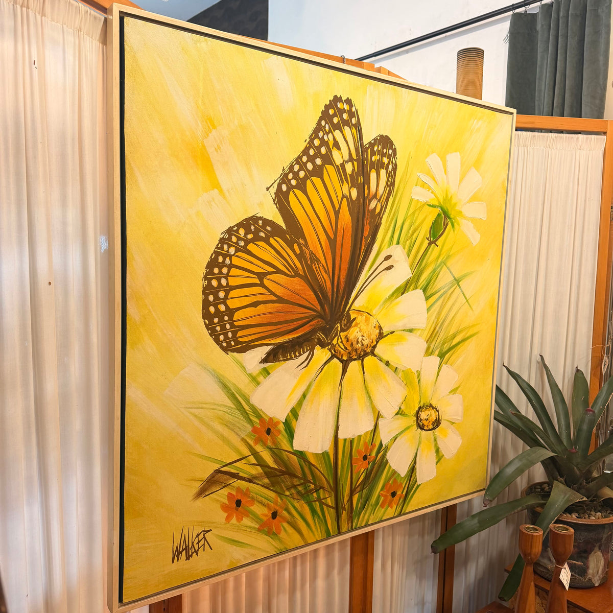 A large framed canvas painting featuring an orange and black butterfly perched on white flowers against a bright yellow background, displayed indoors on a wooden stand with curtains and a potted plant nearby.