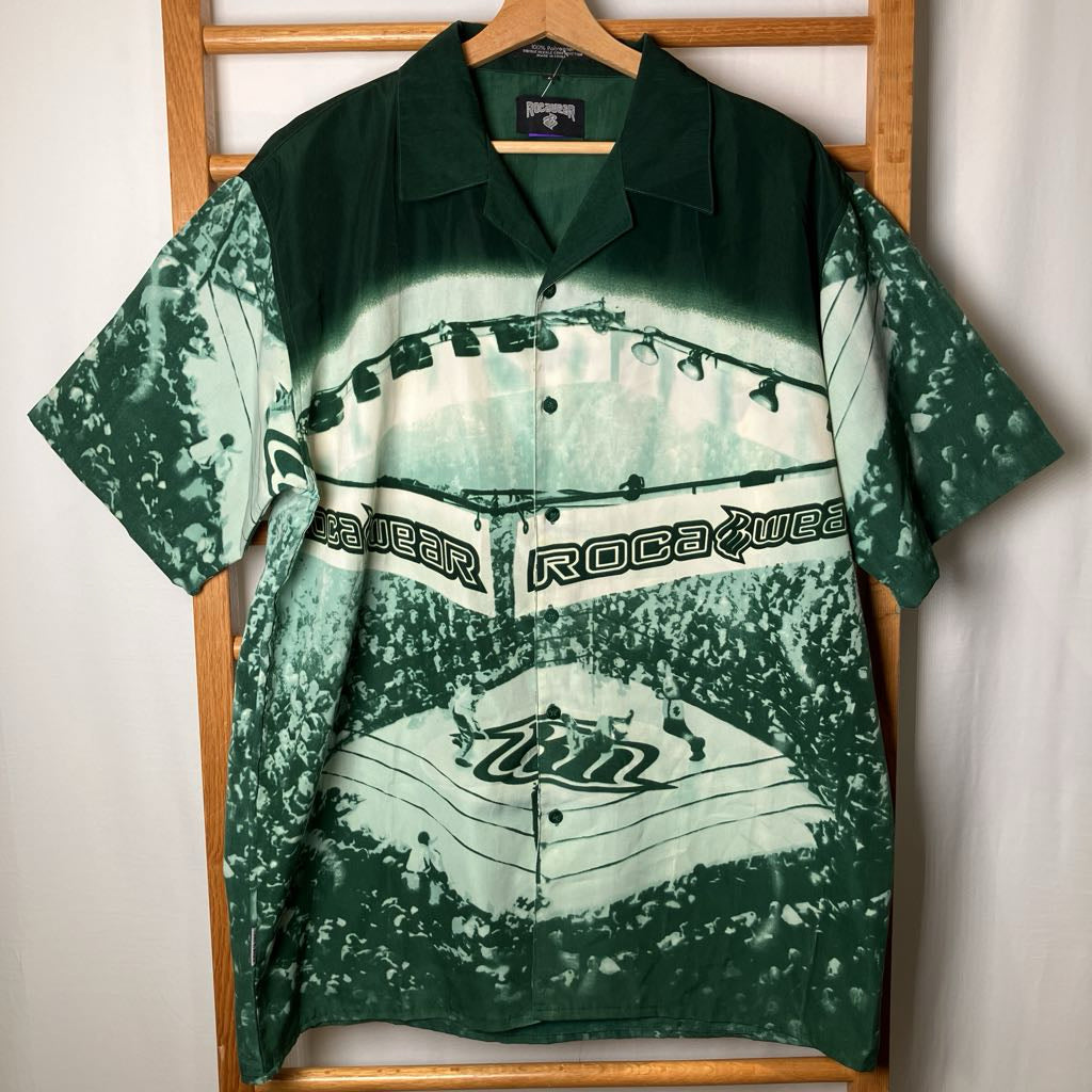 A short-sleeve button-up camp shirt with a green and white tie-dye baseball stadium graphic, hanging on a wooden hanger against a wooden ladder.