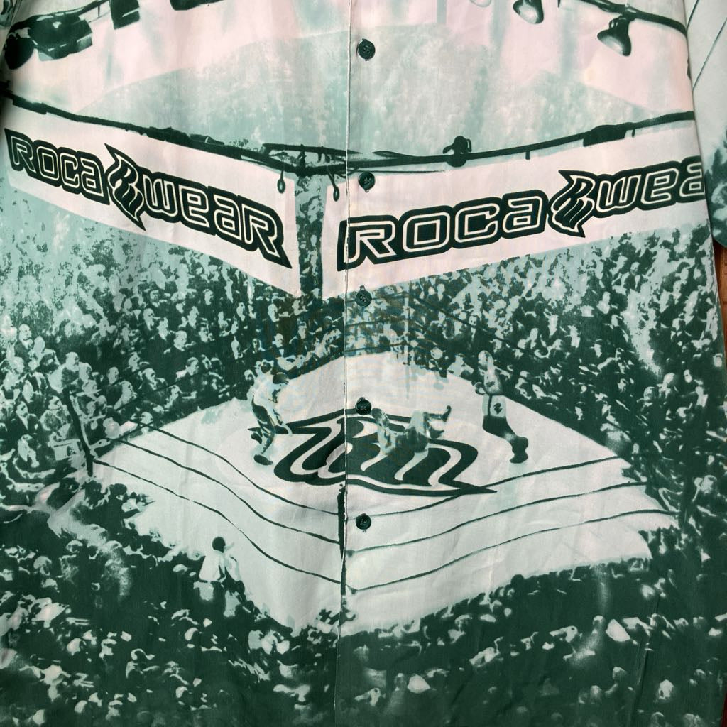 A close-up of a button-front shirt or towel featuring a green, white, and black graphic print with the words 'Roca Wear' across banners and a large 'Win' logo in the center.