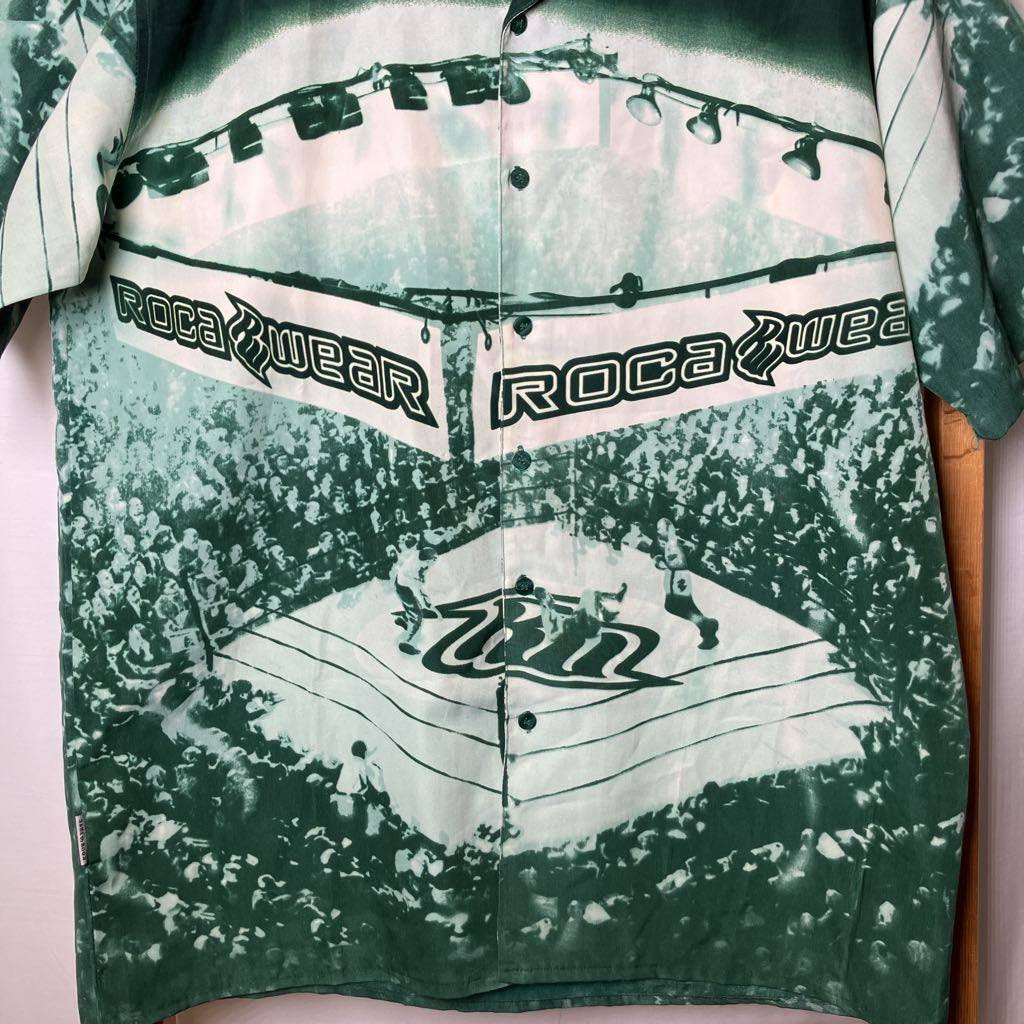 A short-sleeve button-up shirt with a green and white tie-dye rock concert graphic, featuring the text 'ROCA WEAR' across the chest.
