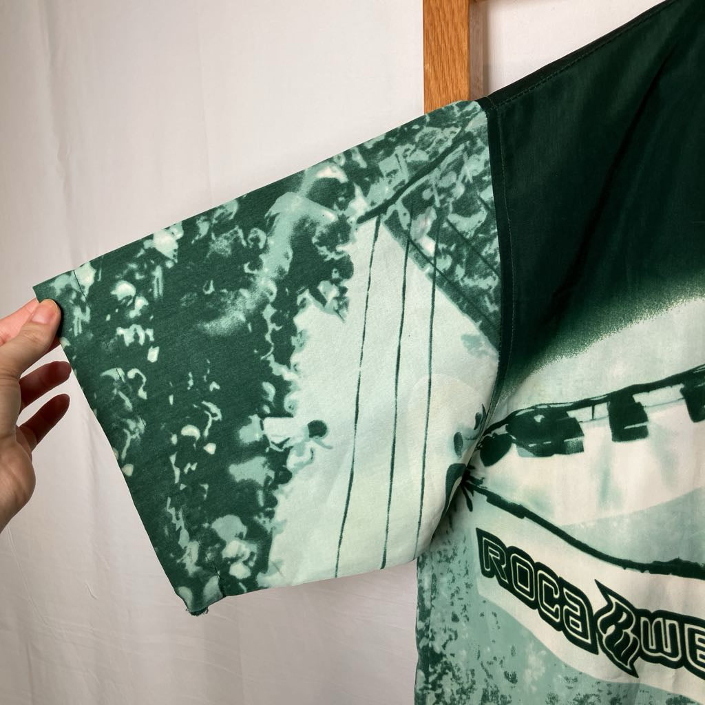 A green and white tie-dye short-sleeve shirt with a graphic print, held up by a hand against a white fabric backdrop.