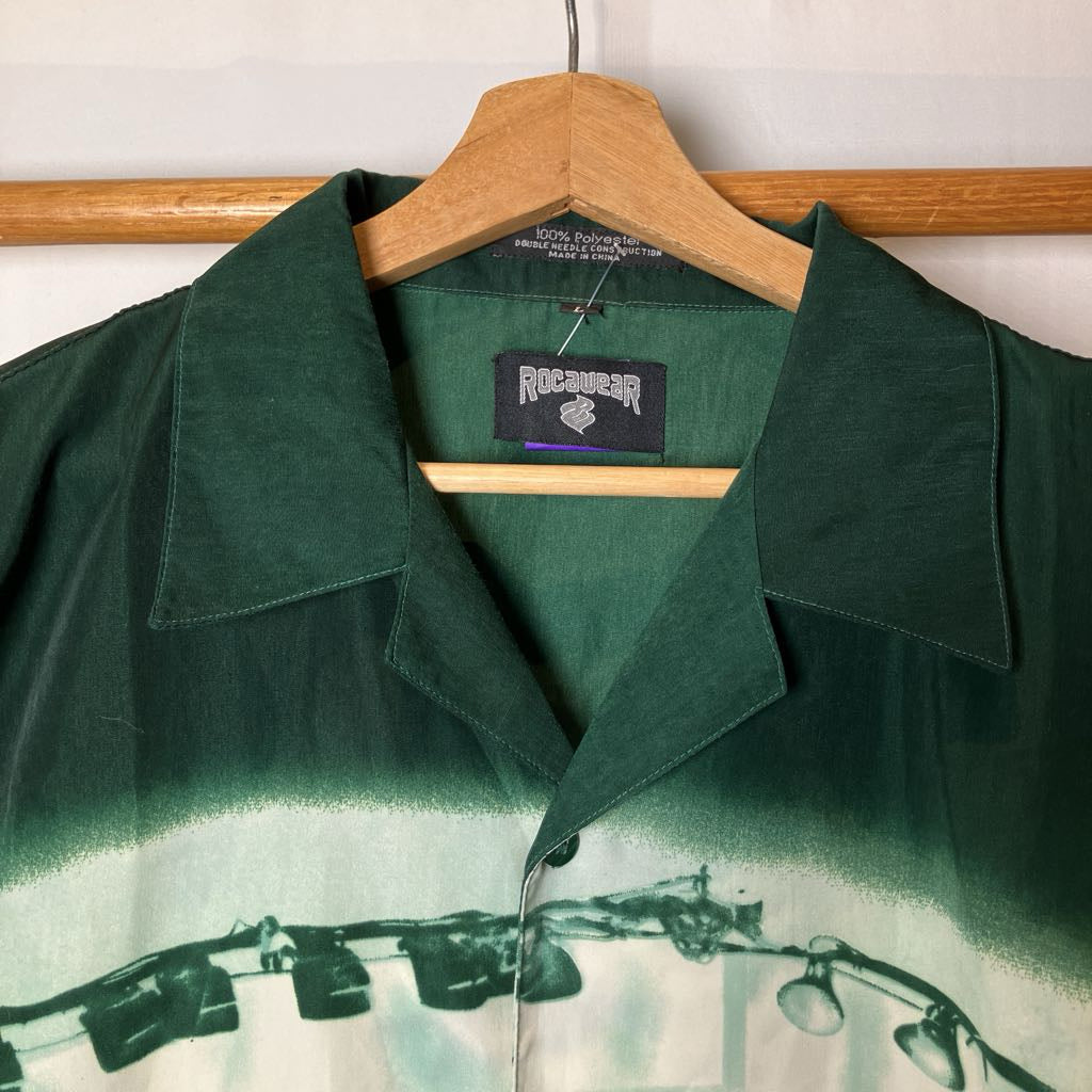 A dark green collared shirt jacket with a gradient fade and a black-and-white photography equipment graphic at the bottom, hanging on a wooden hanger against a white wall.