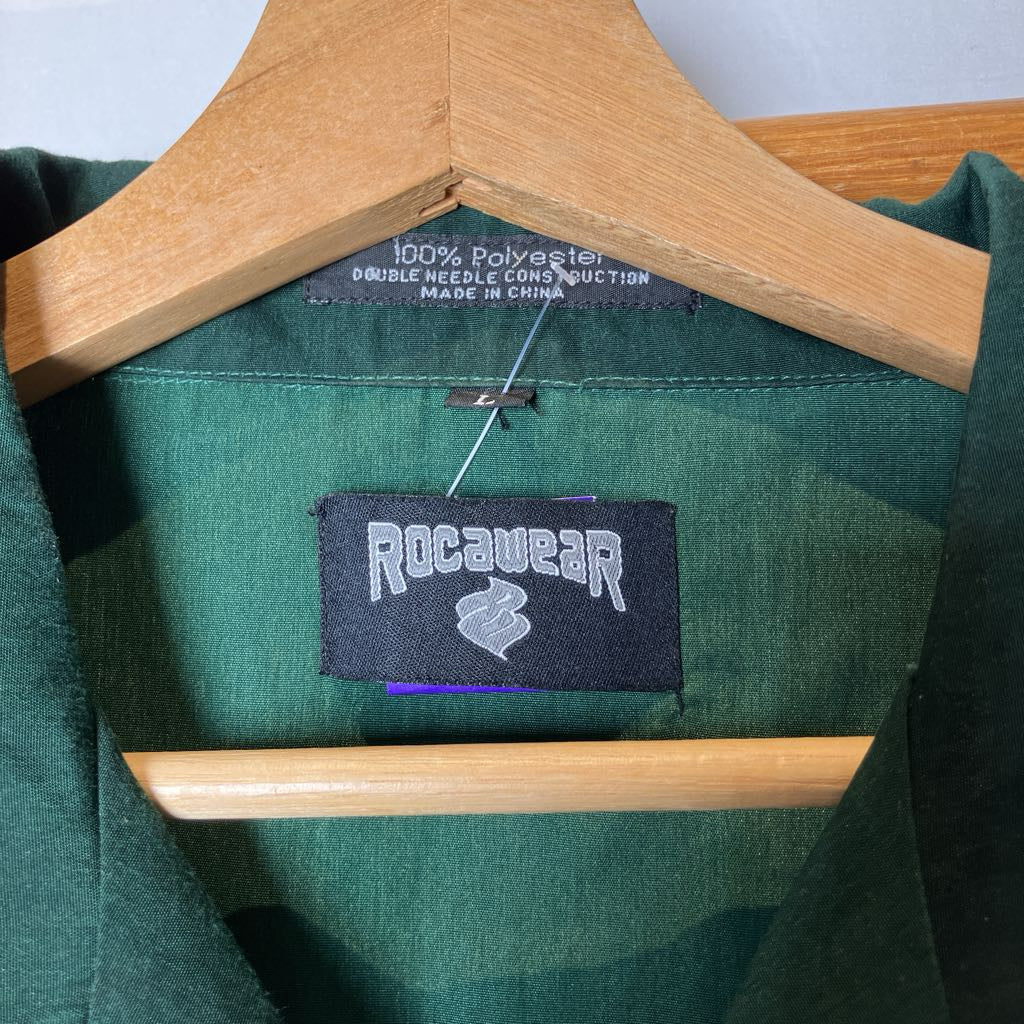 A close-up of a dark green garment hanging on a wooden hanger, showing the interior neck label and a sewn-on brand patch that reads 'Rocawear'.
