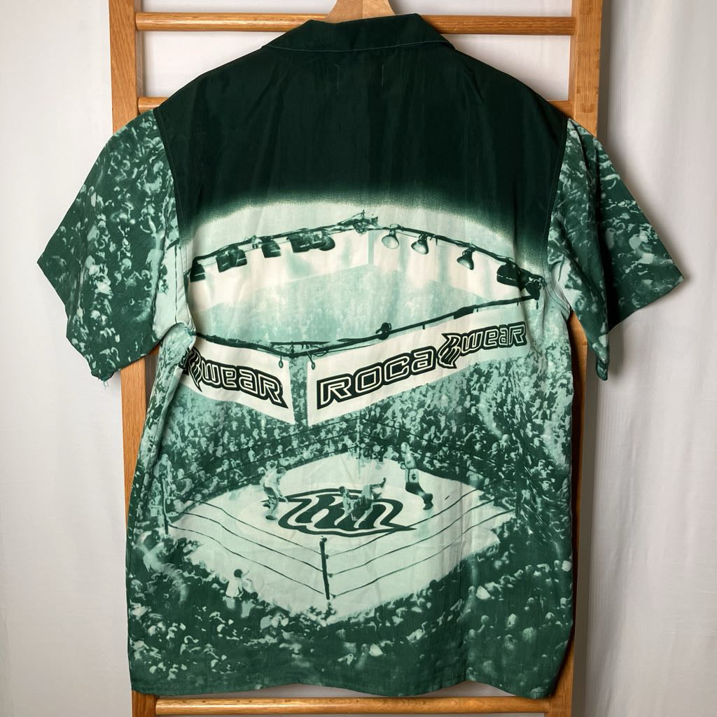 A green short-sleeve button-up shirt with a large green-and-white graphic print of a boxing ring and crowd, hanging on a wooden hanger against a white wall.
