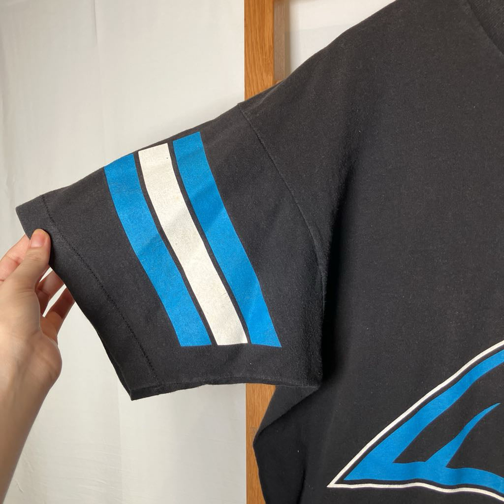A close-up of a black short-sleeve t-shirt being held by a hand, showing a sleeve graphic with two vertical stripes in bright blue and white, bordered by thin black lines.