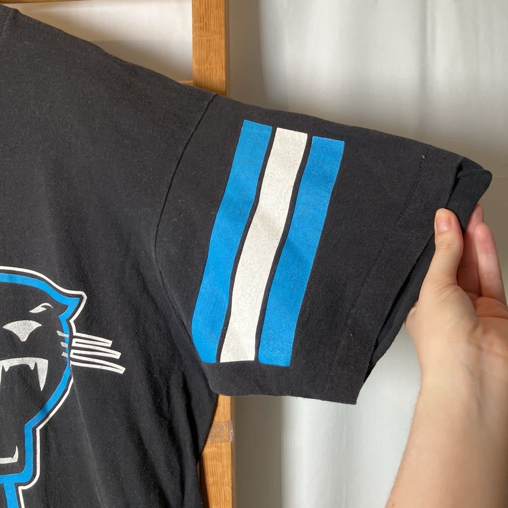 A close-up of a black Carolina Panthers t-shirt being held by a hand, showing the sleeve with blue and white stripes and part of the panther logo.