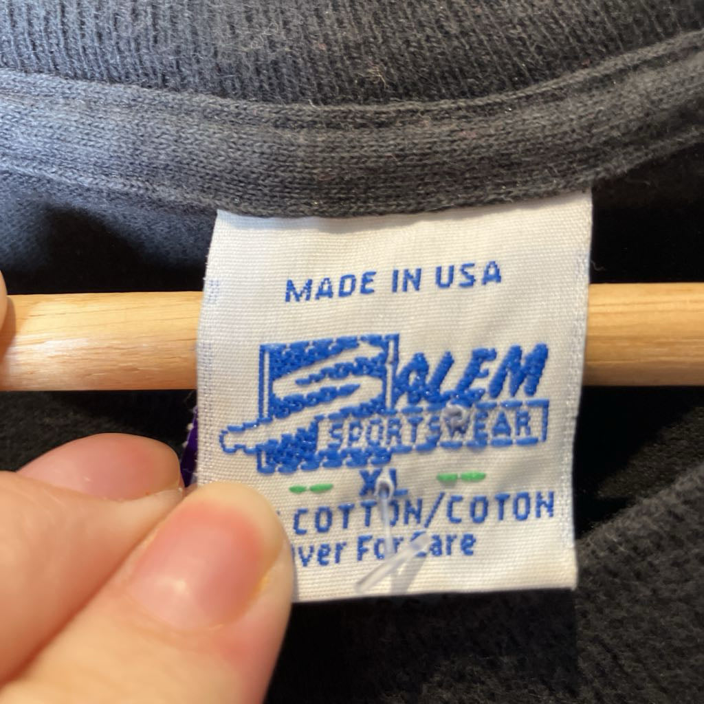 A close-up of a clothing label reading 'Salem Sports Wear' and 'MADE IN USA', held by a hand near a wooden hanger against dark gray knit fabric.