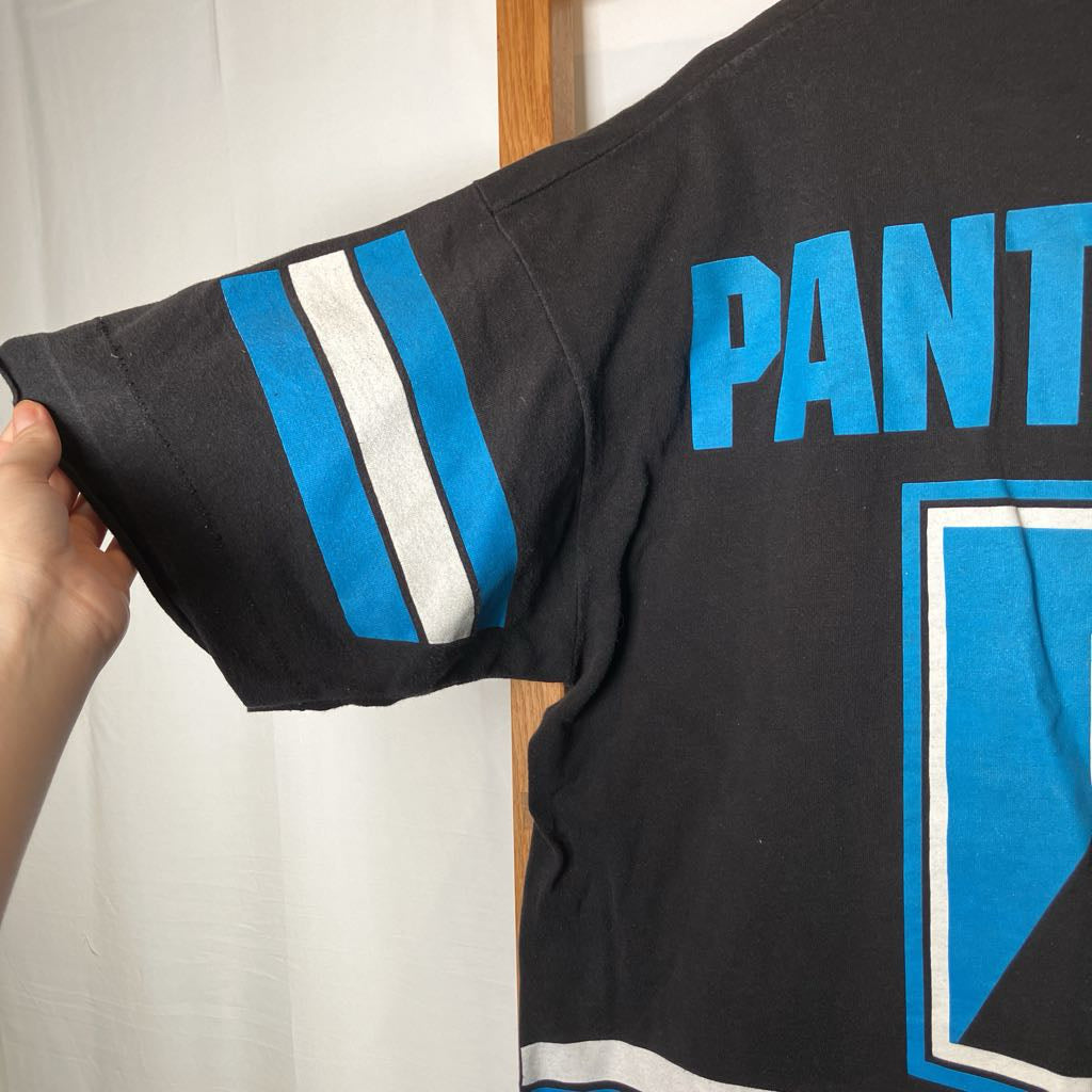 A close-up of a black short-sleeve jersey being held by a hand, showing blue and white sleeve stripes and the partial blue text 'PANT' on the back.