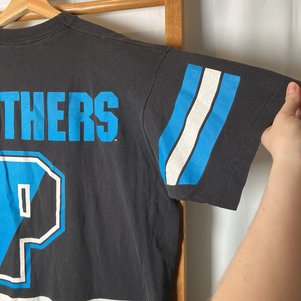 A black vintage athletic t-shirt with blue and white graphics, hanging on a wooden hanger with a hand holding the sleeve.