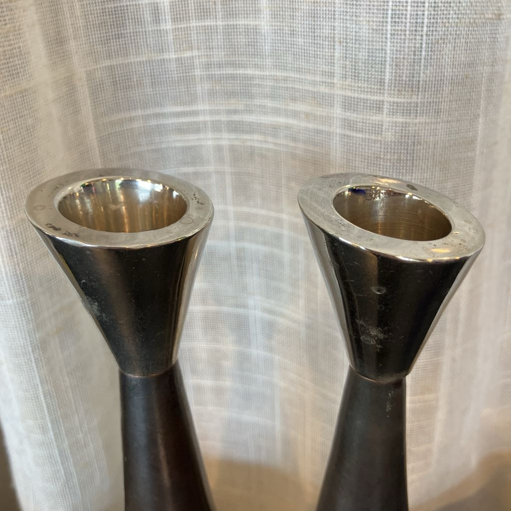 Two dark metal candlestick holders with flared silver tops, shown against a white textured curtain background.