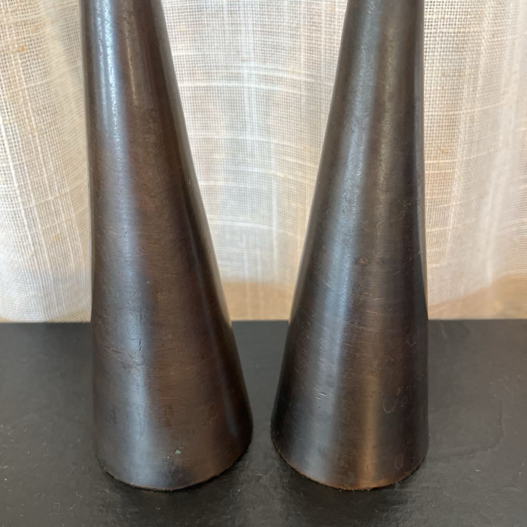 A pair of dark brown, cone-shaped decorative objects with a brushed metal or wood texture, standing upright on a dark surface in front of a light beige curtain.