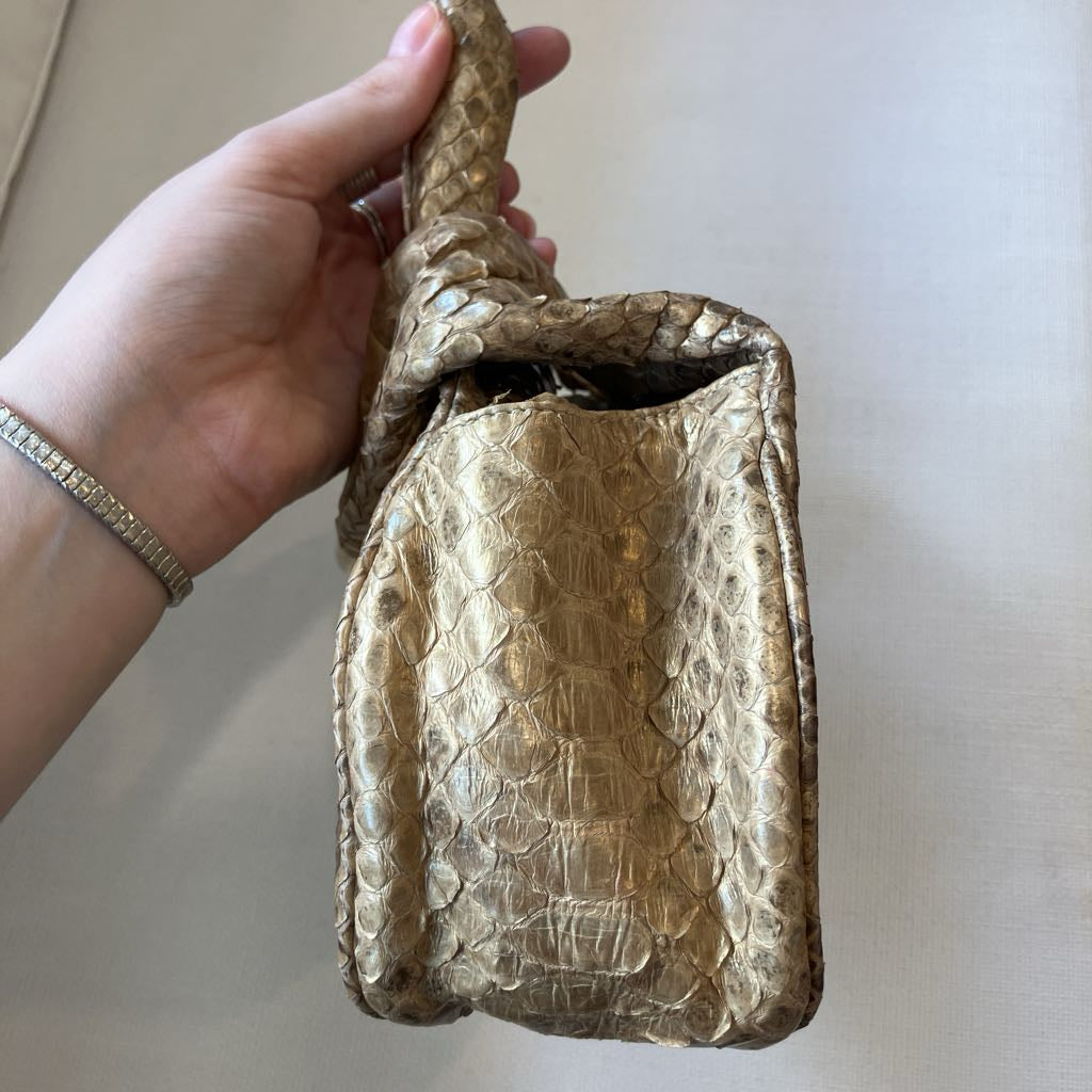 A hand holding a small metallic gold handbag with a textured python skin pattern and a woven handle, set against a neutral beige background.
