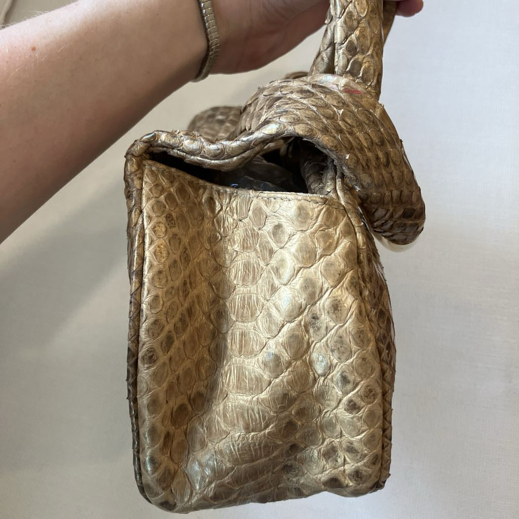 A close-up of a hand holding a small structured handbag made from textured natural reptile skin, showing a metallic gold and brown alligator or crocodile pattern against a plain light background.