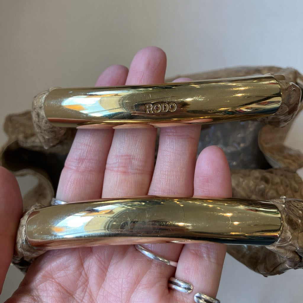 A close-up of a hand holding two polished gold-tone metal handles, with one handle showing the engraved brand name 'RODO'. The handles are wrapped in protective brown paper on the ends, and the background is a soft beige wall.