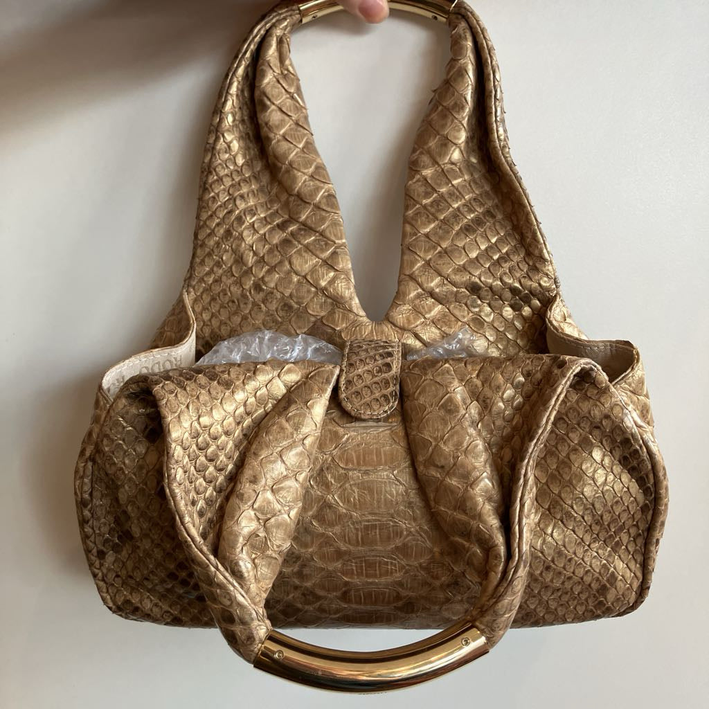 A metallic gold textured shoulder bag with a snakeskin pattern, featuring a curved gold metal handle and a small top button closure, shown against a plain off-white background.