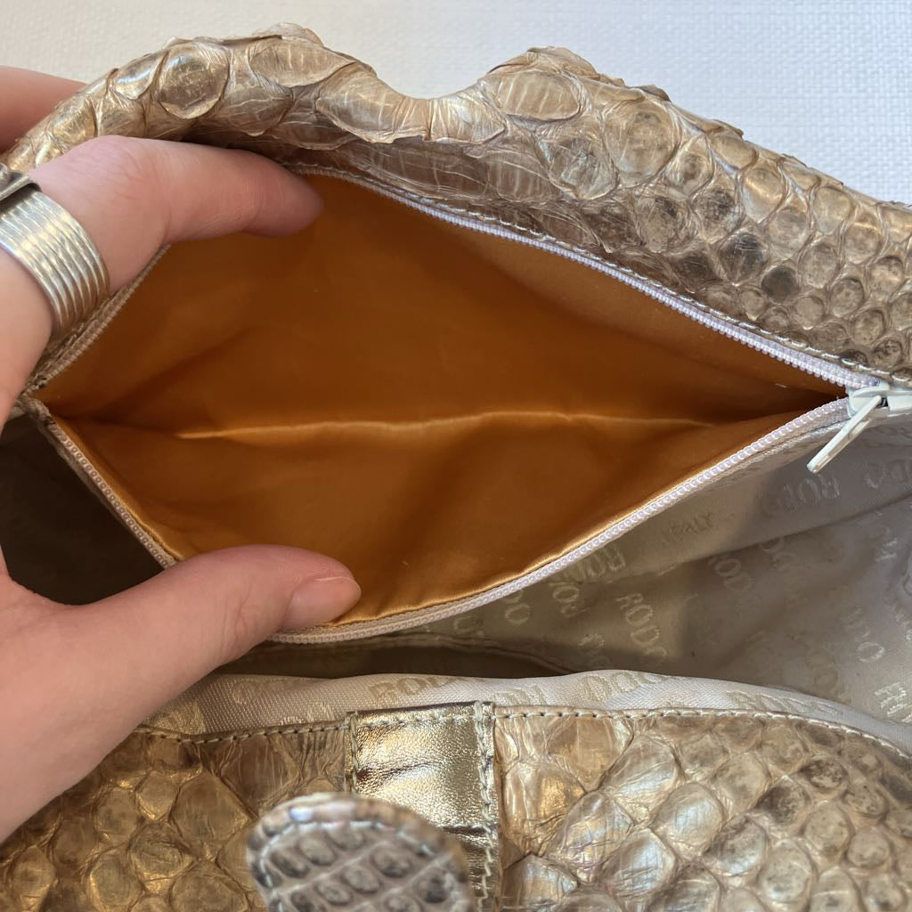 Close-up of a metallic gold and beige python-textured clutch bag being held open by a hand, showing a gold satin lining and a zipper closure.