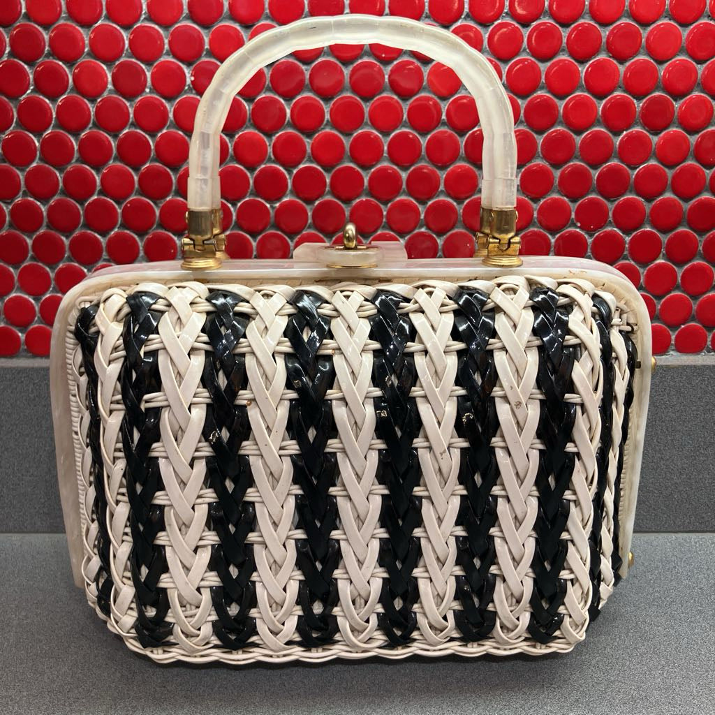 A structured handbag with a black and off-white woven body, a clear plastic top handle, and gold-tone hardware, set against a red circular tile background.