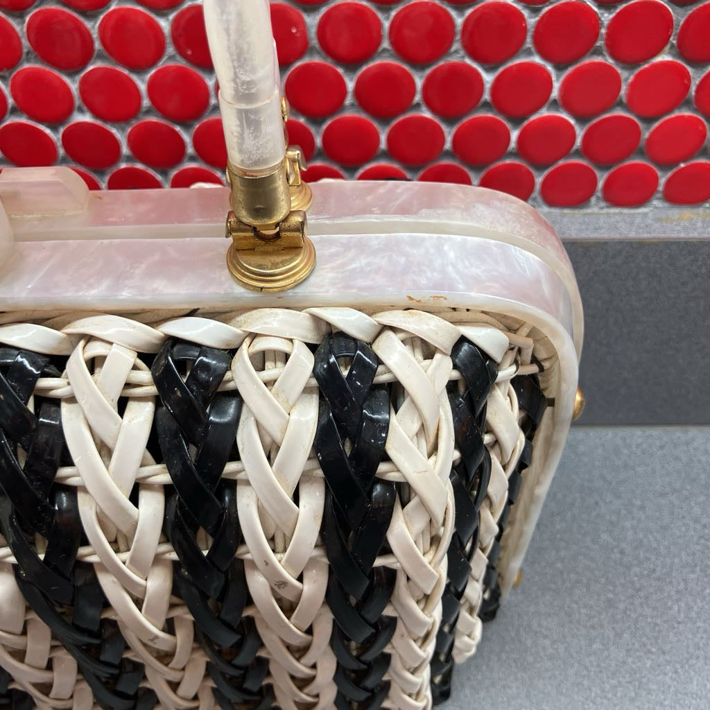 Close-up of a woven clutch handbag with black and cream leather straps, a translucent pink shell, and a gold clasp, set against a red mosaic tile background.